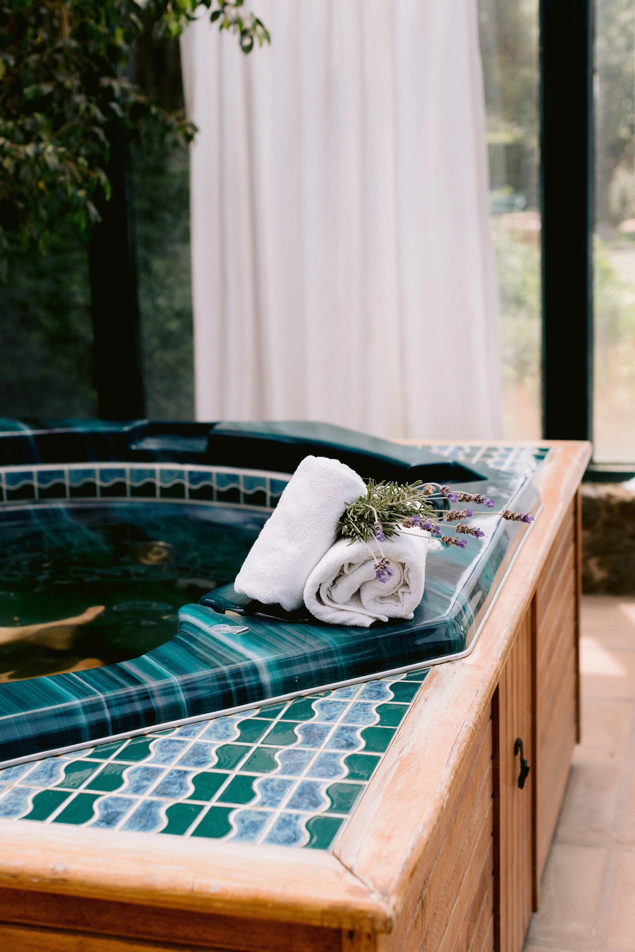 Jacuzzi de madera y azulejos con toallas blancas enrolladas y lavanda, en un entorno luminoso con cortinas blancas.