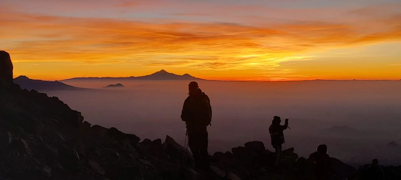 Tres personas en un risco al amanecer, con vista a una capa de nubes y montañas en la distancia