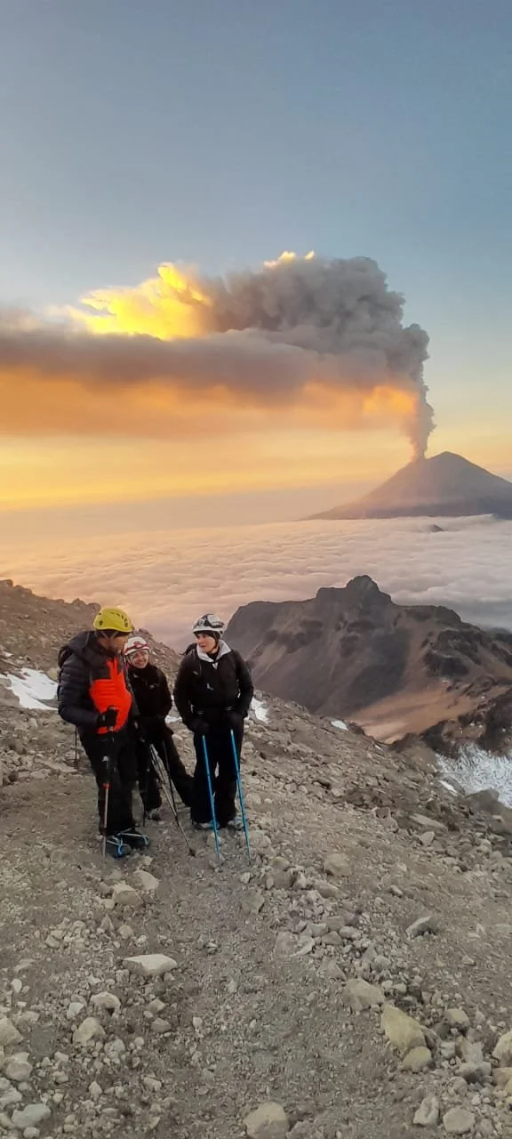Tres personas con equipo de alpinismo en una ruta de montaña, con volcán en erupción en el fondo y cielo al amanecer.