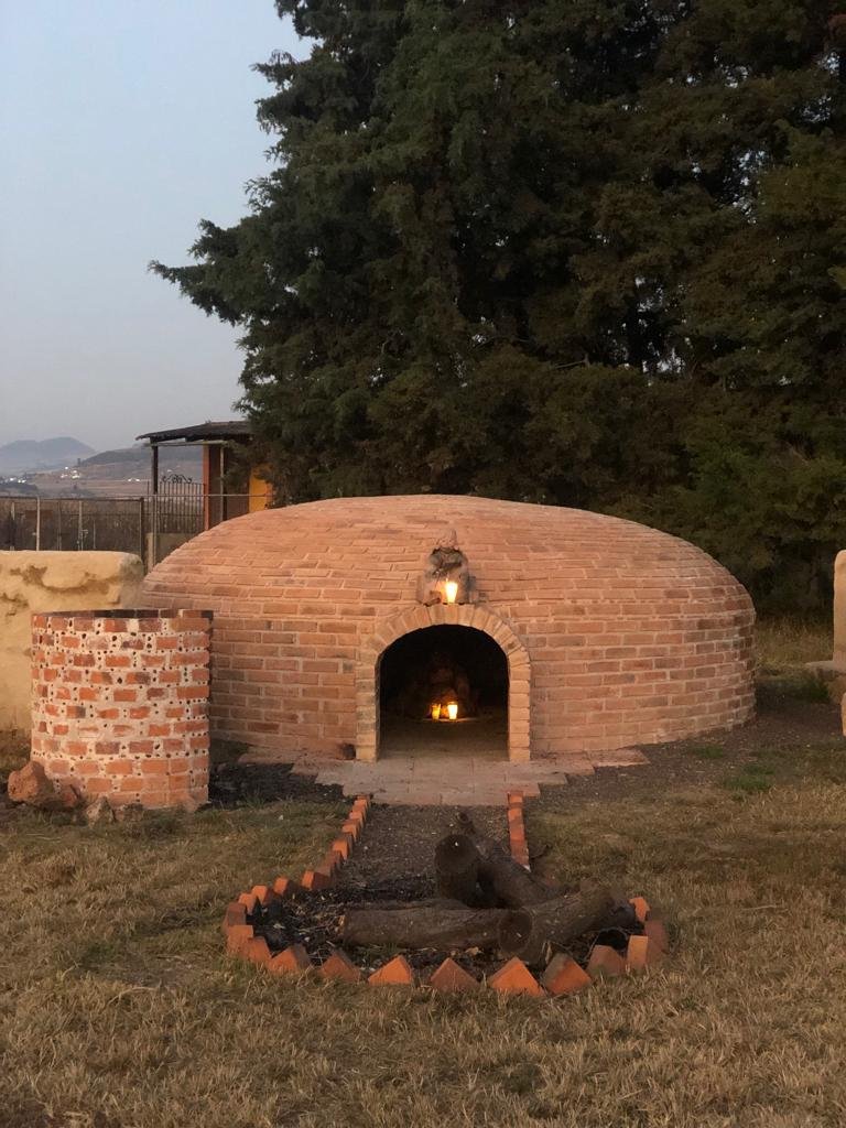 Horno de leña de ladrillo con fogata frente y en su interior, en un área al aire libre con árboles y cielo al atardecer.