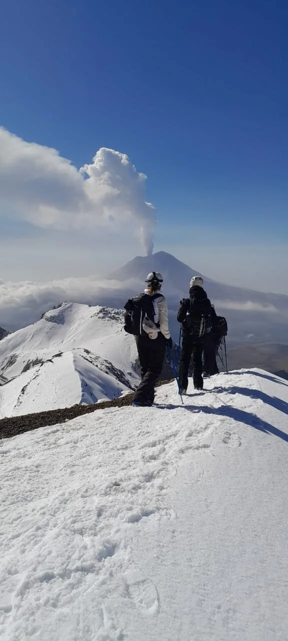 Dos excursionistas con equipo de montaña observando el volcán en erupción desde la nieve, con una columna de humo saliendo del volcán en el fondo.