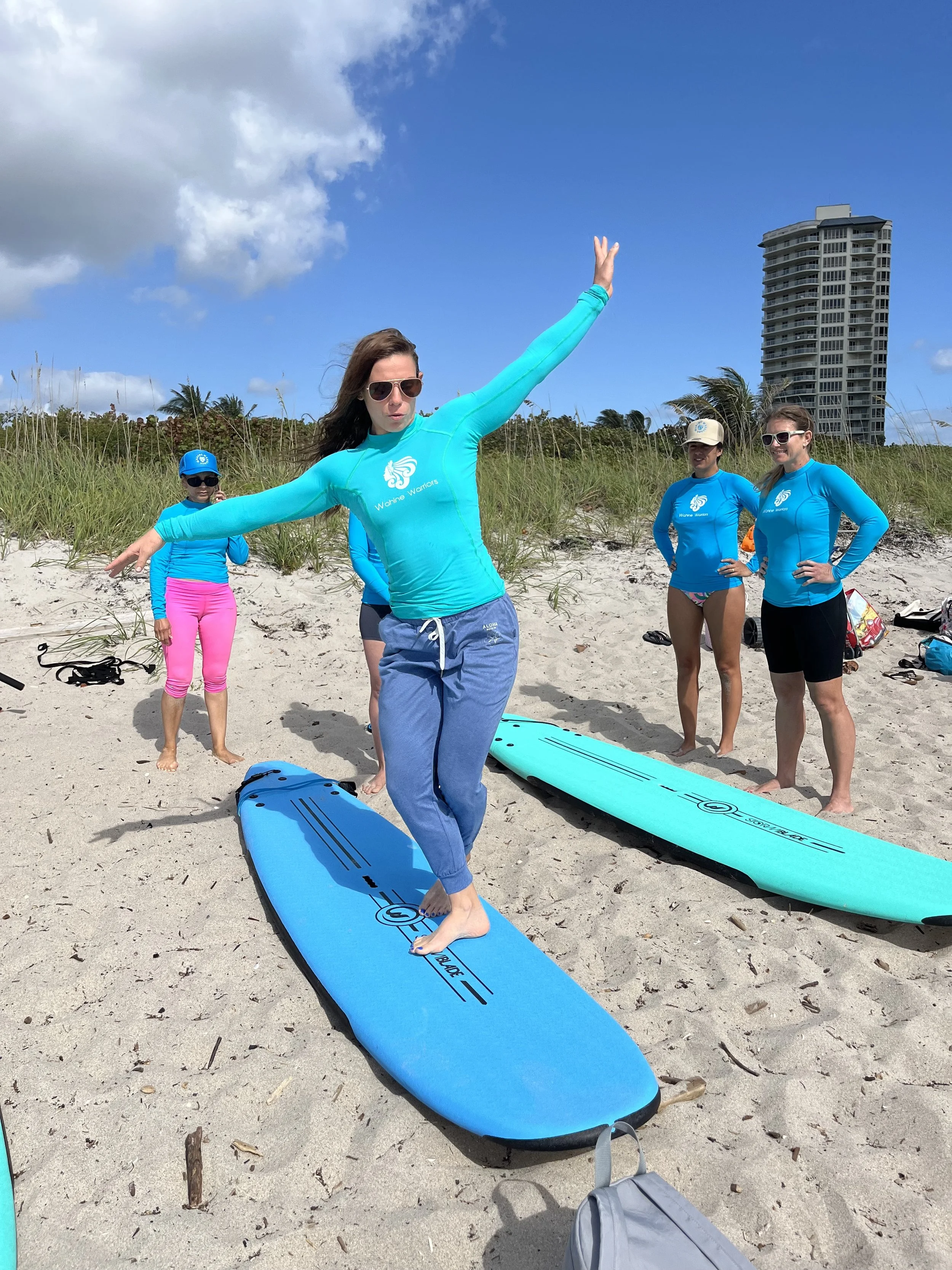 A woman balancing on a paddleboard on the beach while a group of women in blue shirts watch. The background includes sand, grass, and a tall building under a partly cloudy sky.