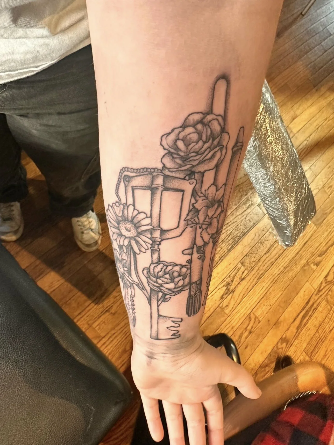 Outline of a tattoo on a person's forearm featuring flowers, a box, and straw brushes.