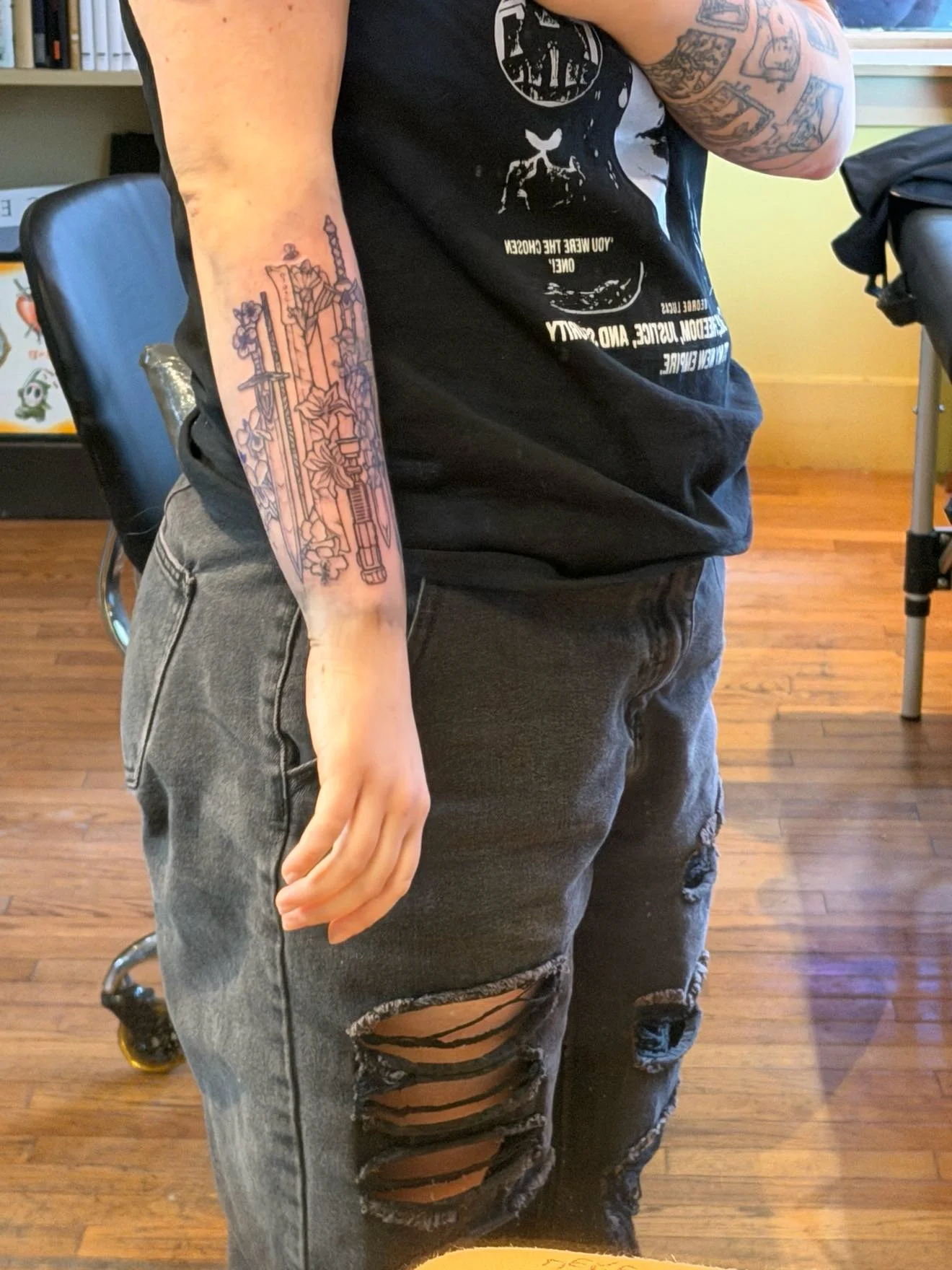 A person with a sleeve tattoo on their left arm and distressed black jeans. They are wearing a black T-shirt with white graphic text and standing in a room with hardwood flooring, a black chair, and a yellow wall.