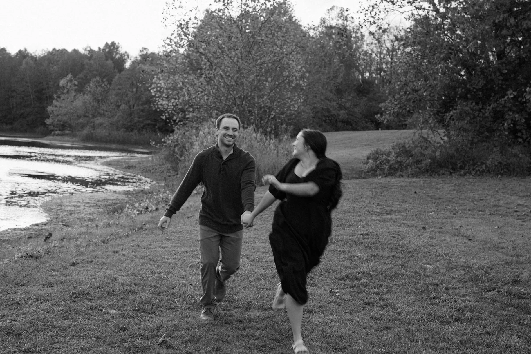 A young couple runs in the field next to a lake at a local park in Morrow Ohio for engagement photos.