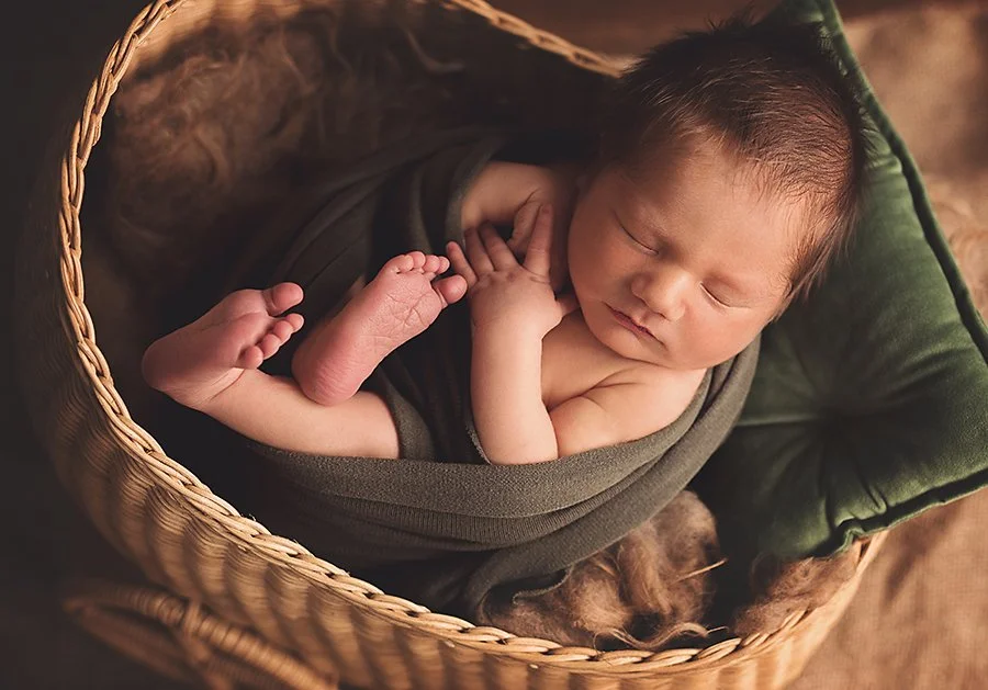Newborn safely posed in a wicker stroller, peacefully sleeping with a blanket and small green pillow under their head.