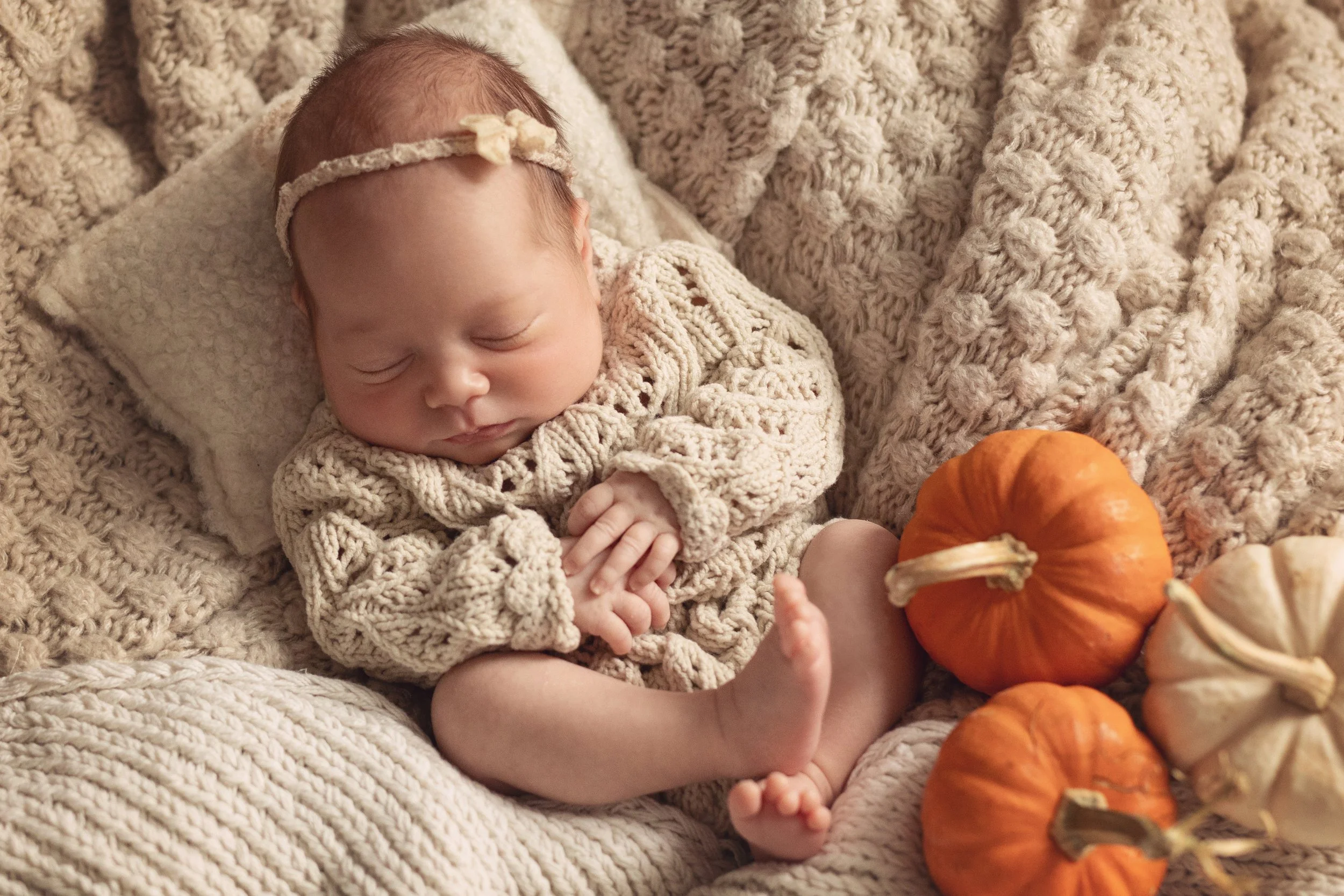 Sleeping newborn in soft window natural light on cream textures in cream lace outfit with pumpkins nestled at feet.