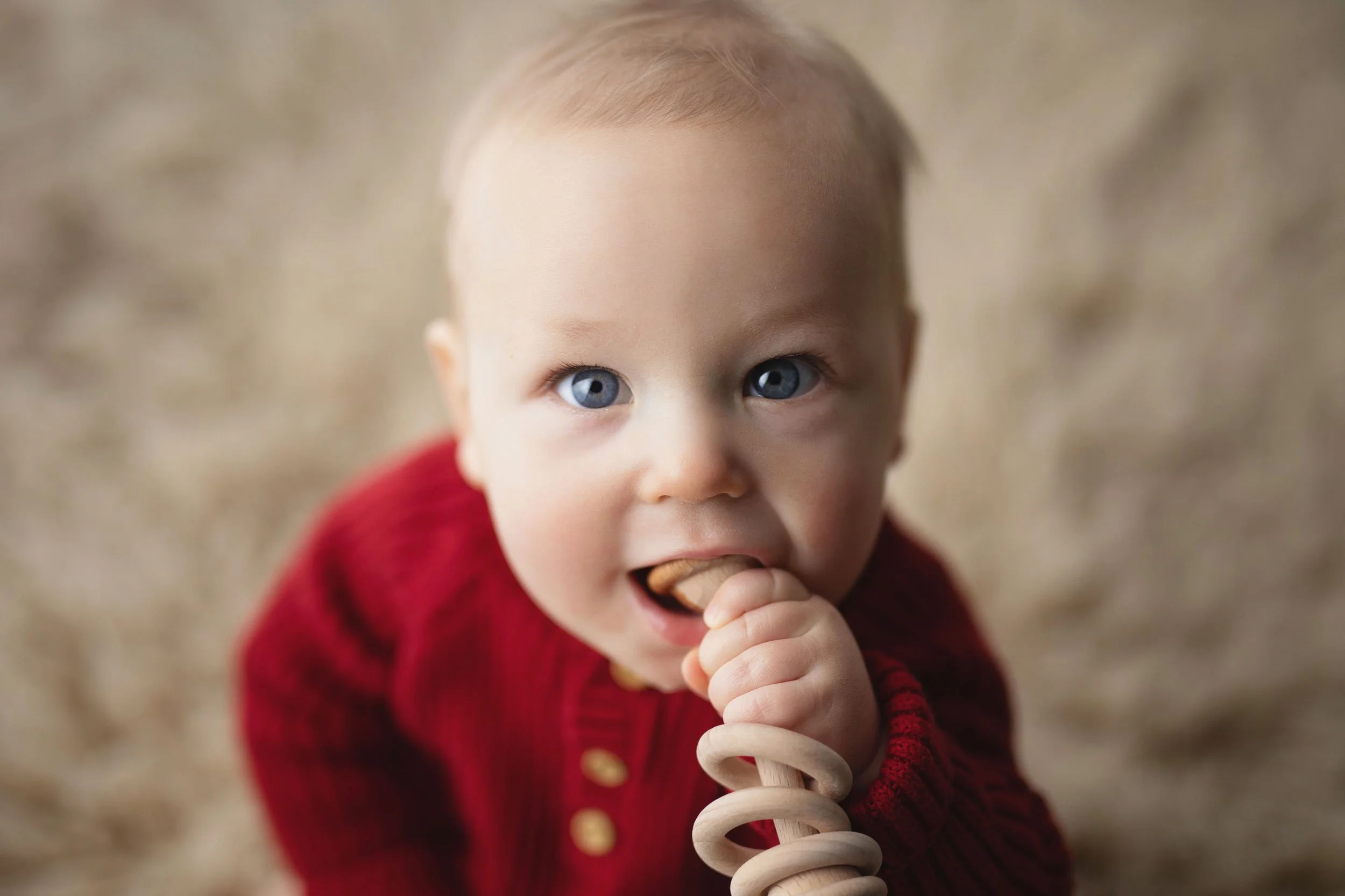 Baby in a red sweater chewing on a wooden spiral teething toy, looking up with bright blue eyes—perfect for BABY MILESTONE PHOTOGRAPHY.