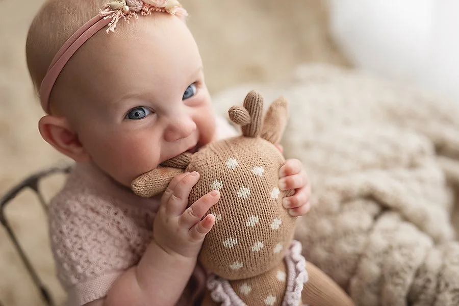 baby girl with blue eyes smiling as she cuddles and teethes on her favorite soft toy from cuddle and kind in a soft window light