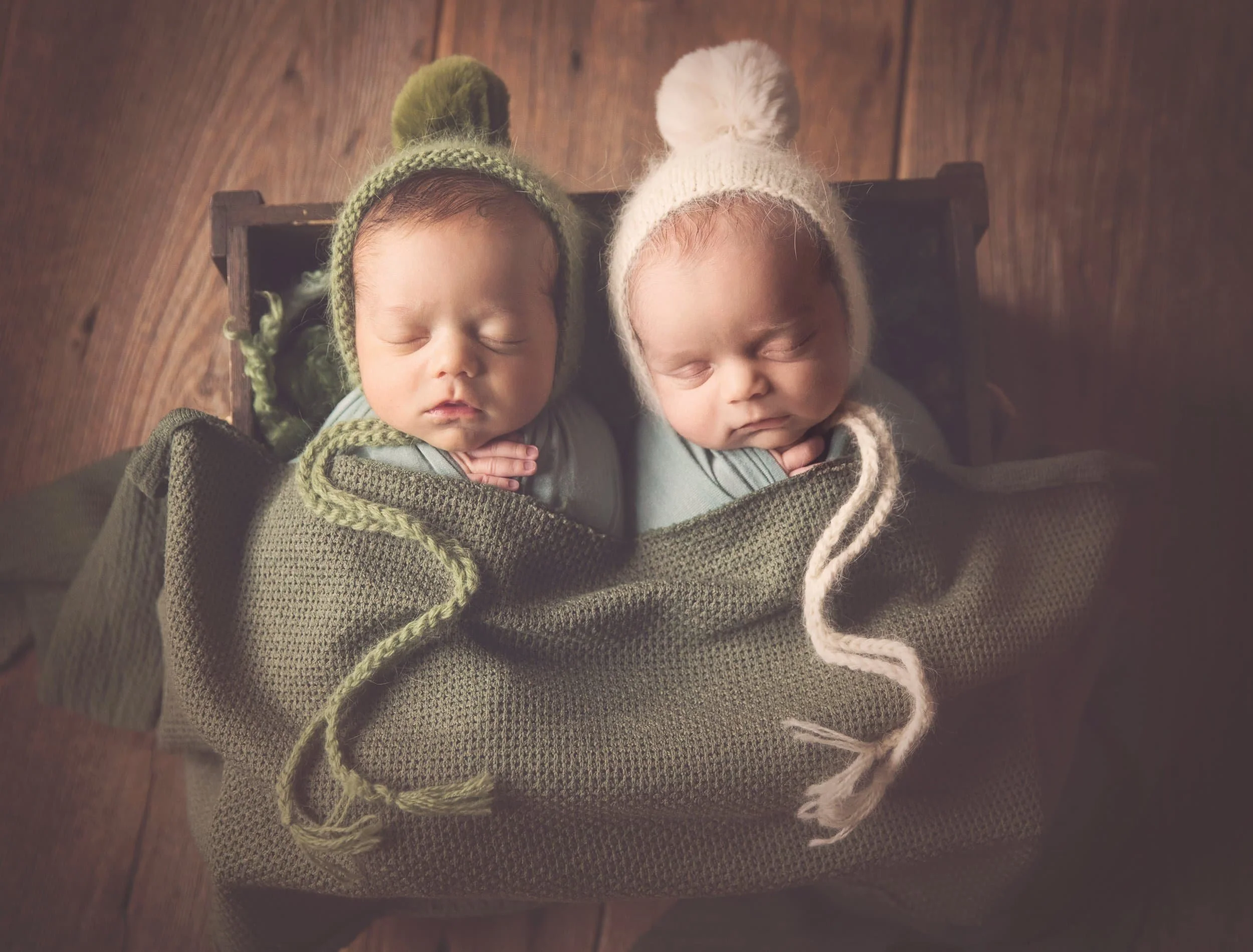 twin newborn babies in pom-pom knit hats swaddled together in crate prop on reclaimed wood studio floor