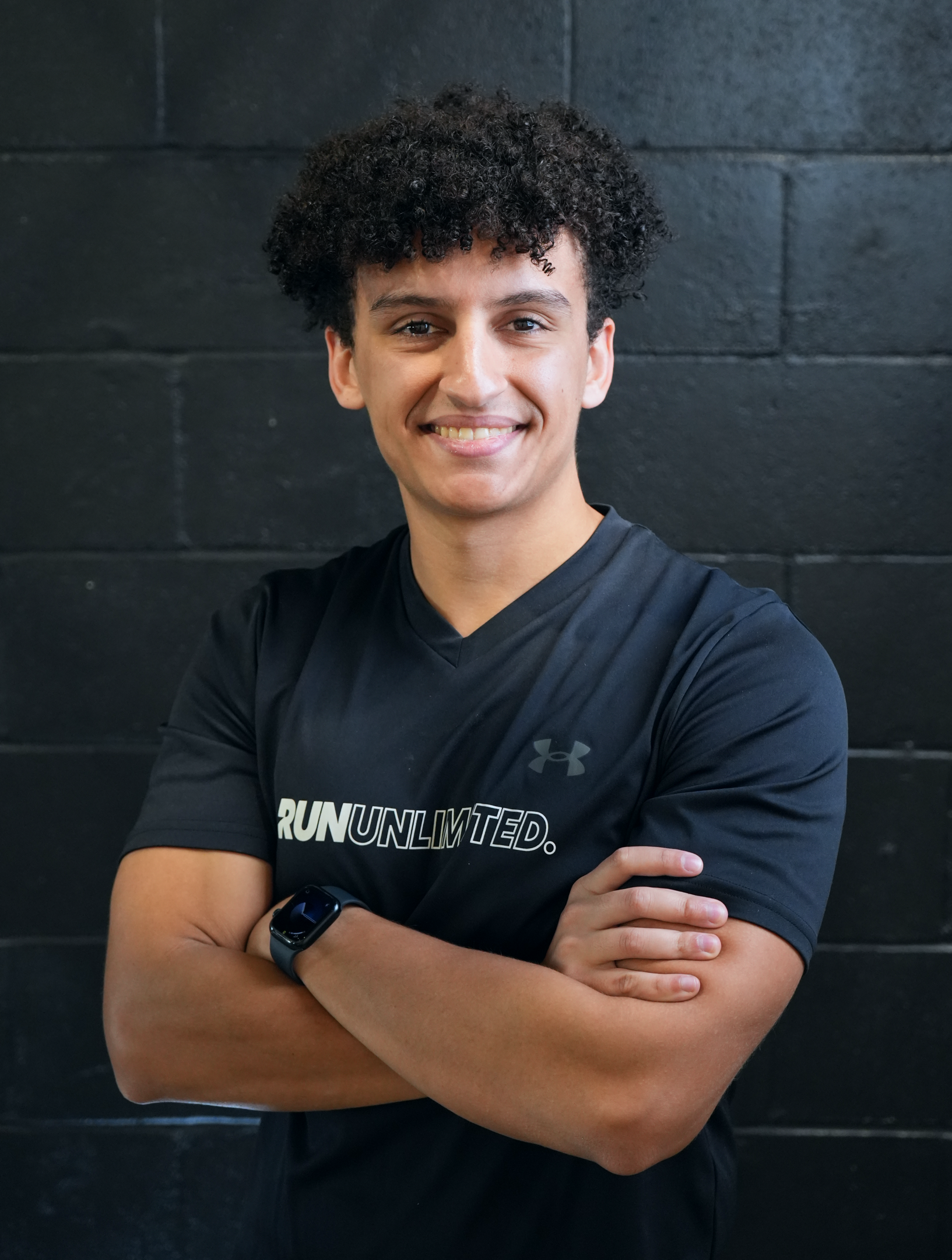 Young man with curly hair smiling with arms crossed, wearing a black athletic shirt with the words 'Run Unlimited' and a sports watch, standing against a dark wall.