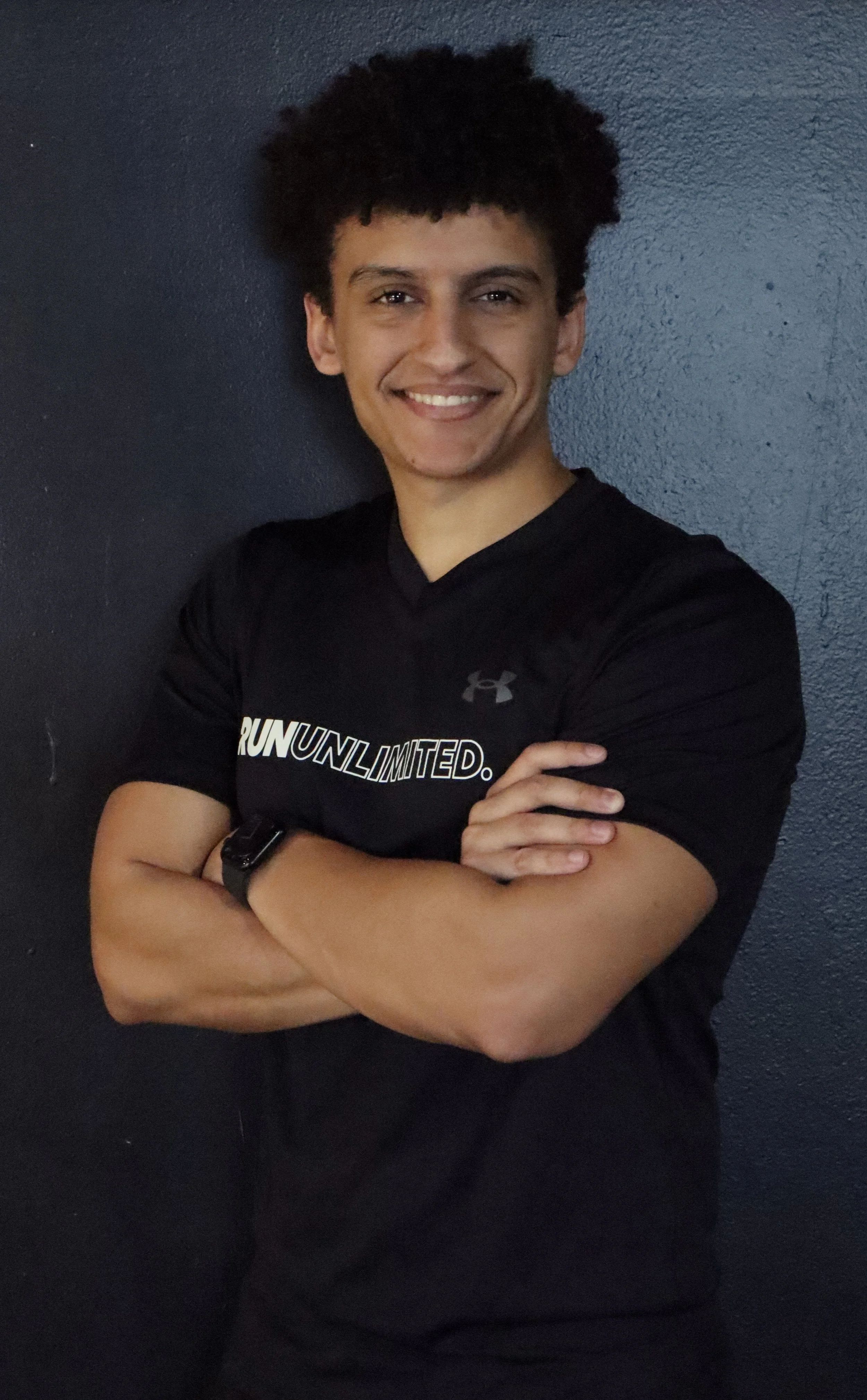 Young man with curly hair smiling with arms crossed, wearing a black athletic shirt with the words 'Run Unlimited' and a sports watch, standing against a dark wall.