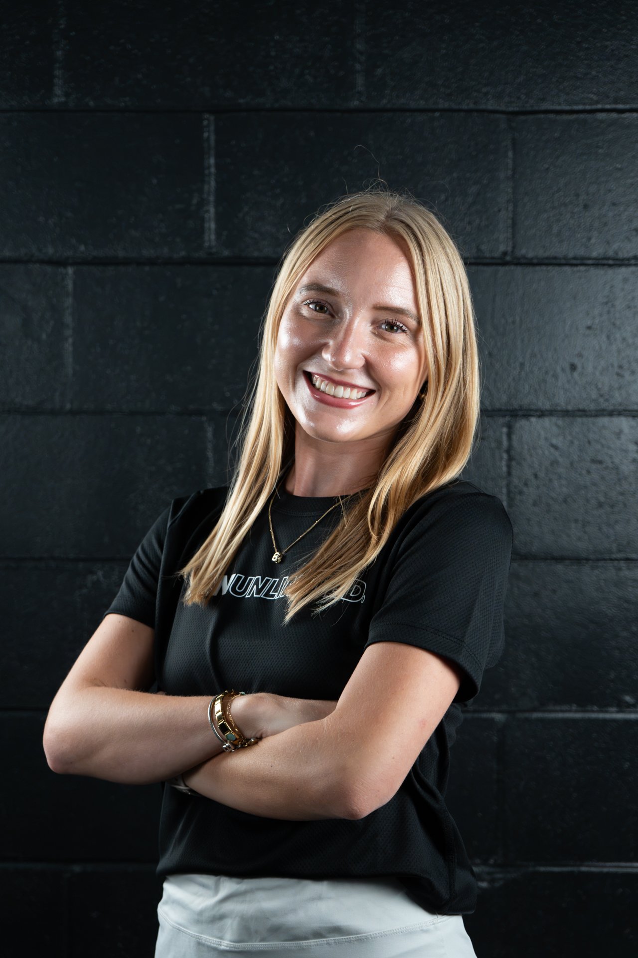 A young woman with long blonde hair, smiling and standing with arms crossed, in front of a black brick wall.