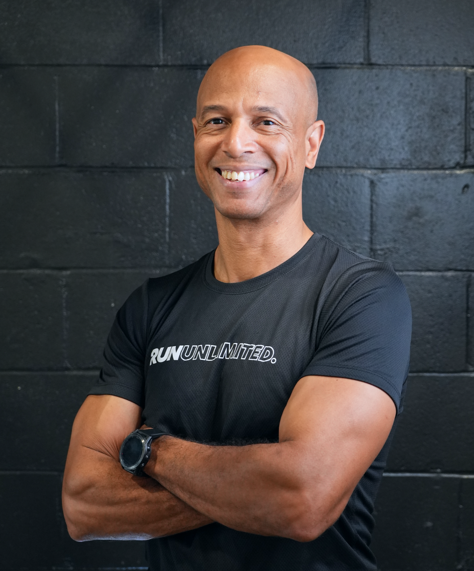 Smiling man wearing a "Run Unlimited" Under Armour shirt, standing against a black brick wall.