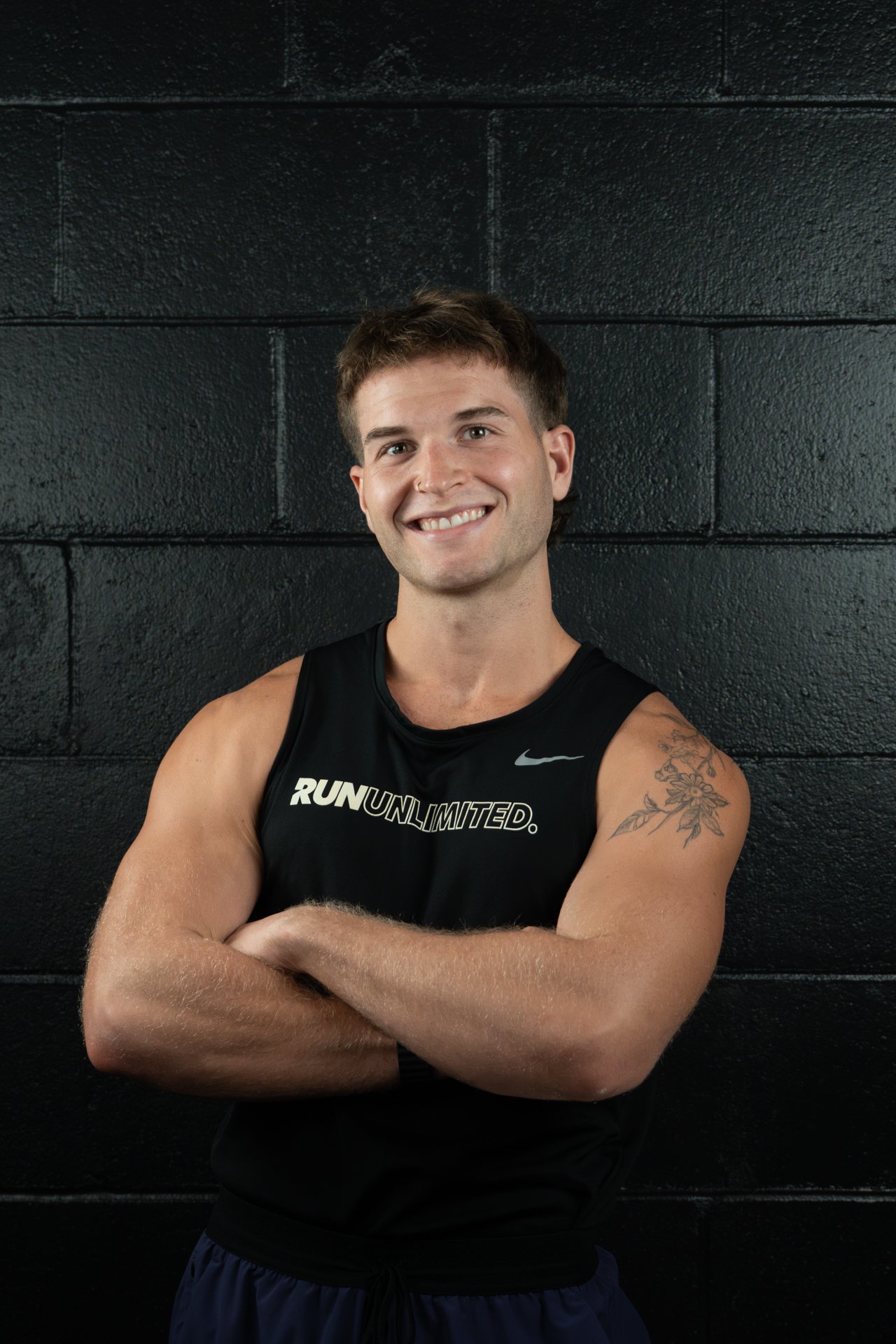 A young man with short brown hair and a nose piercing, smiling with arms crossed, wearing a black sleeveless athletic shirt and a gold chain, standing against a black brick wall.