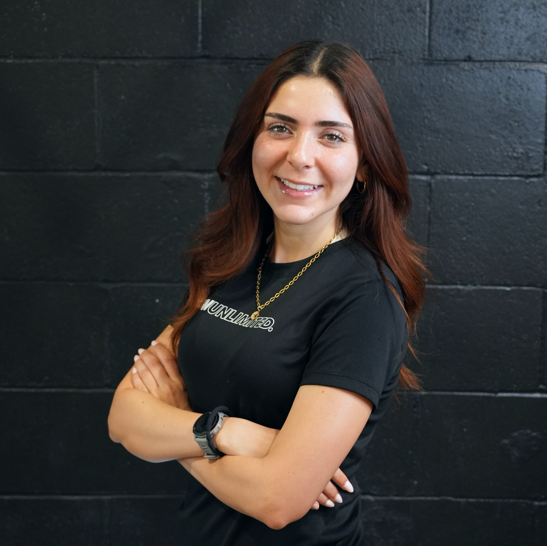 A person with long brown hair and glasses wearing a black t-shirt with the word "UNLIMITED" and crossing their arms, smiling in front of a dark brick wall.