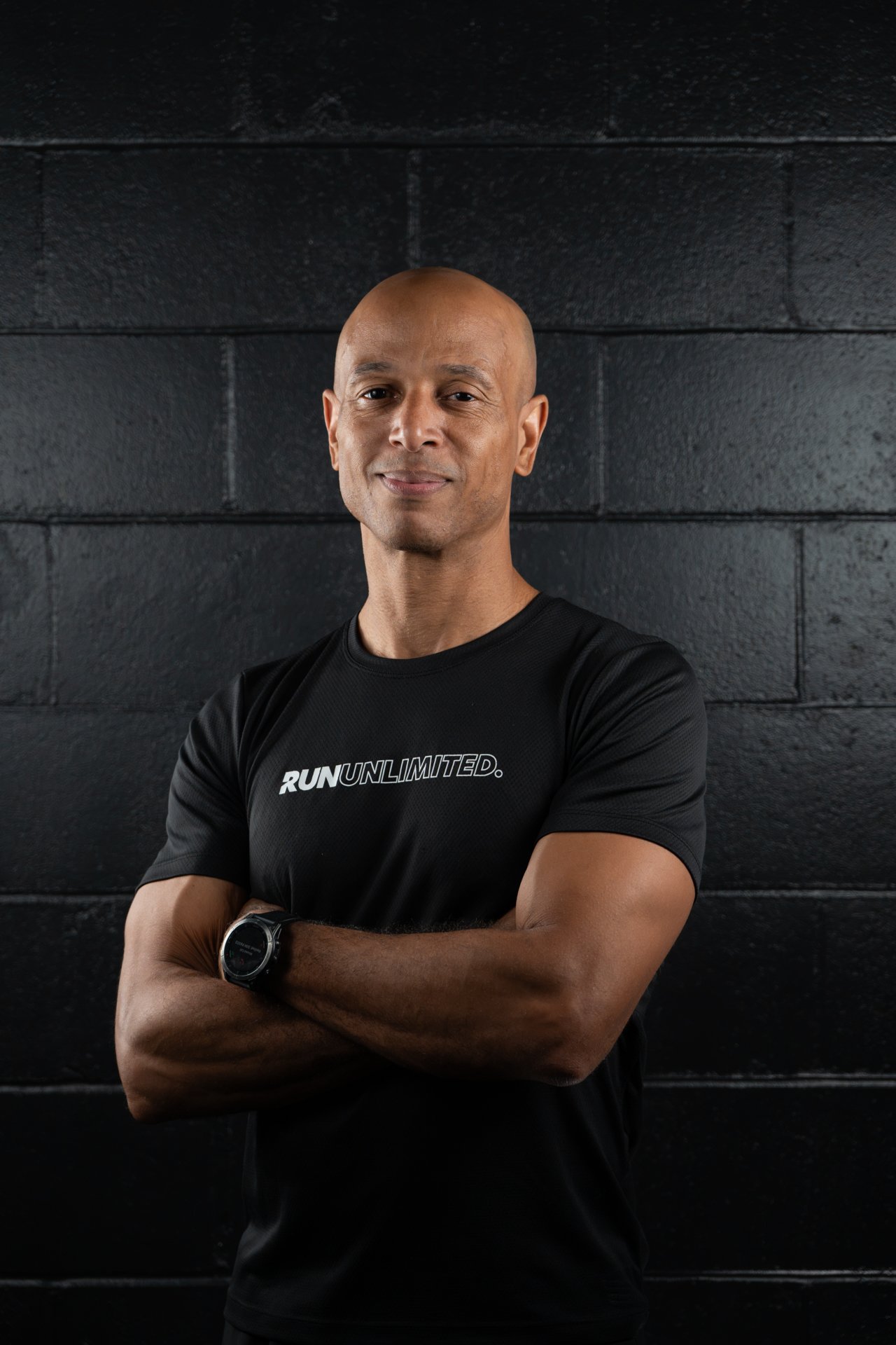 Smiling man wearing a "Run Unlimited" Under Armour shirt, standing against a black brick wall.