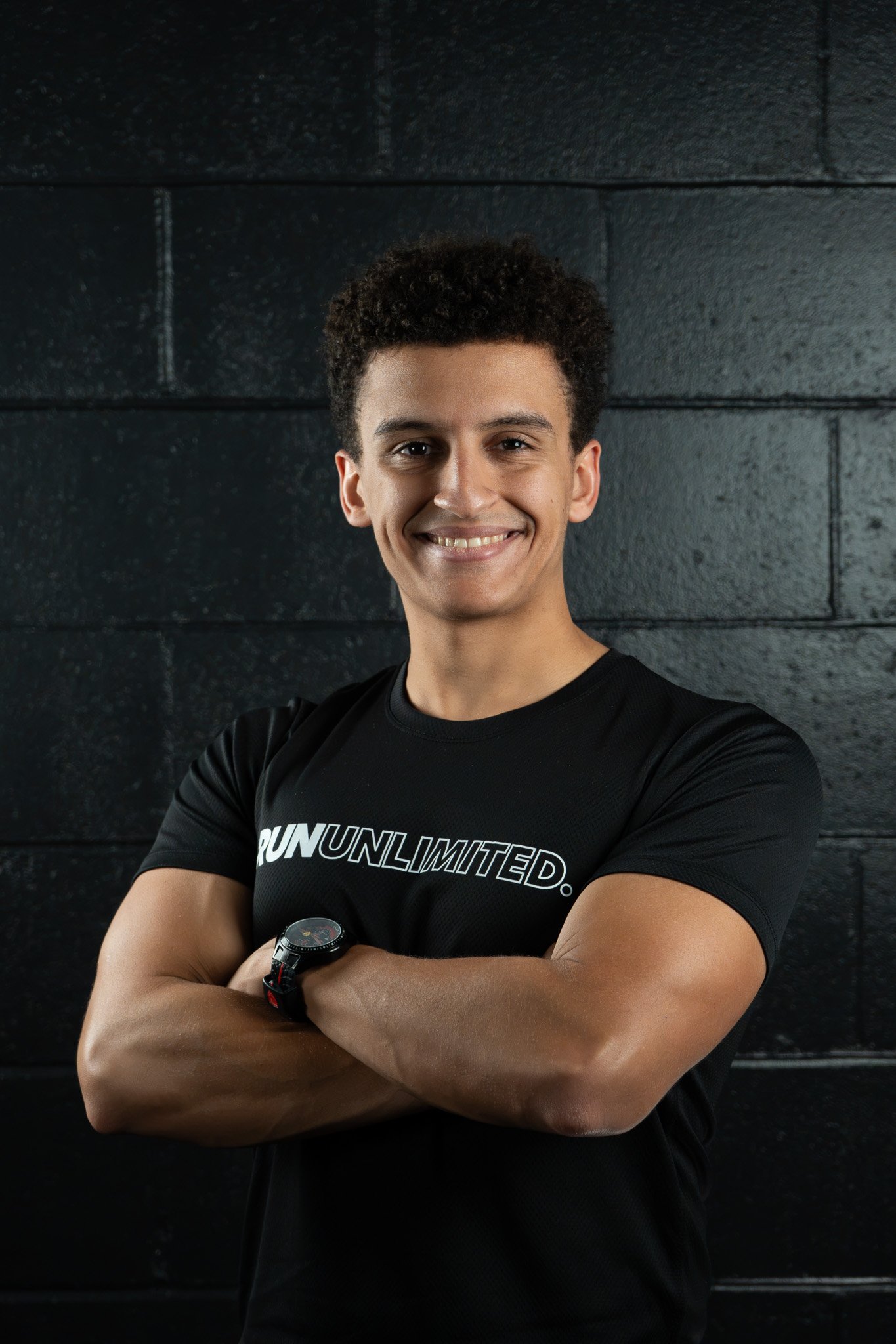 Young man with curly hair smiling with arms crossed, wearing a black athletic shirt with the words 'Run Unlimited' and a sports watch, standing against a dark wall.