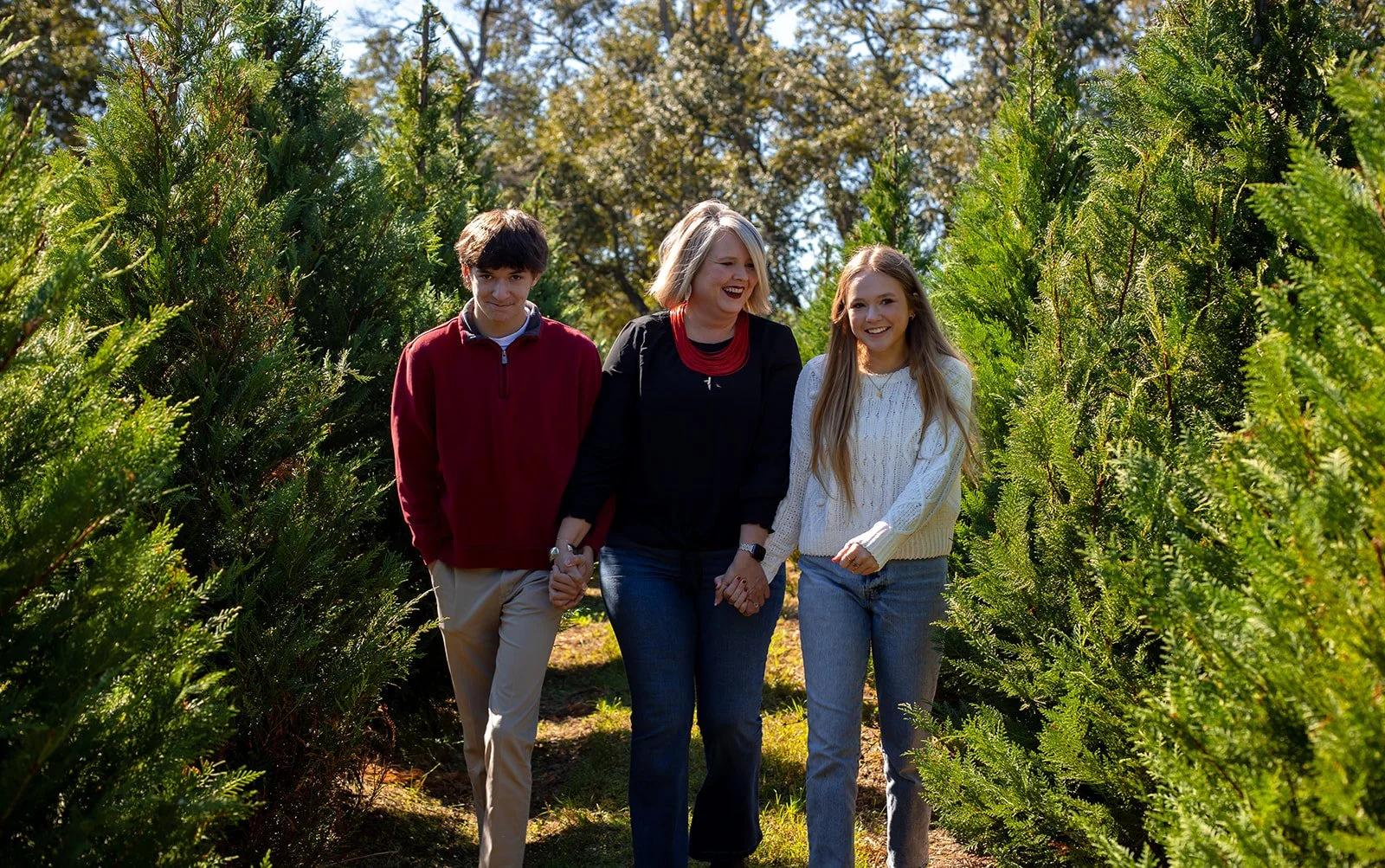 Three people, a woman and two younger individuals, walking hand in hand through a row of evergreen trees outdoors on a sunny day. Richmond, Virginia Christmas Session