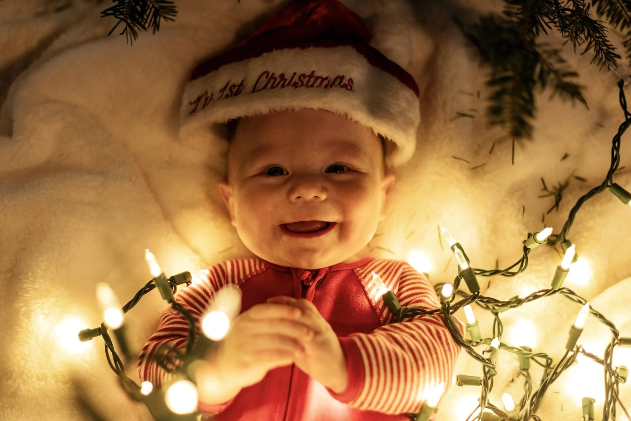 A smiling baby lying on a soft surface, wearing a Santa hat with the words "My 1st Christmas" embroidered on it, surrounded by Christmas lights and greenery.