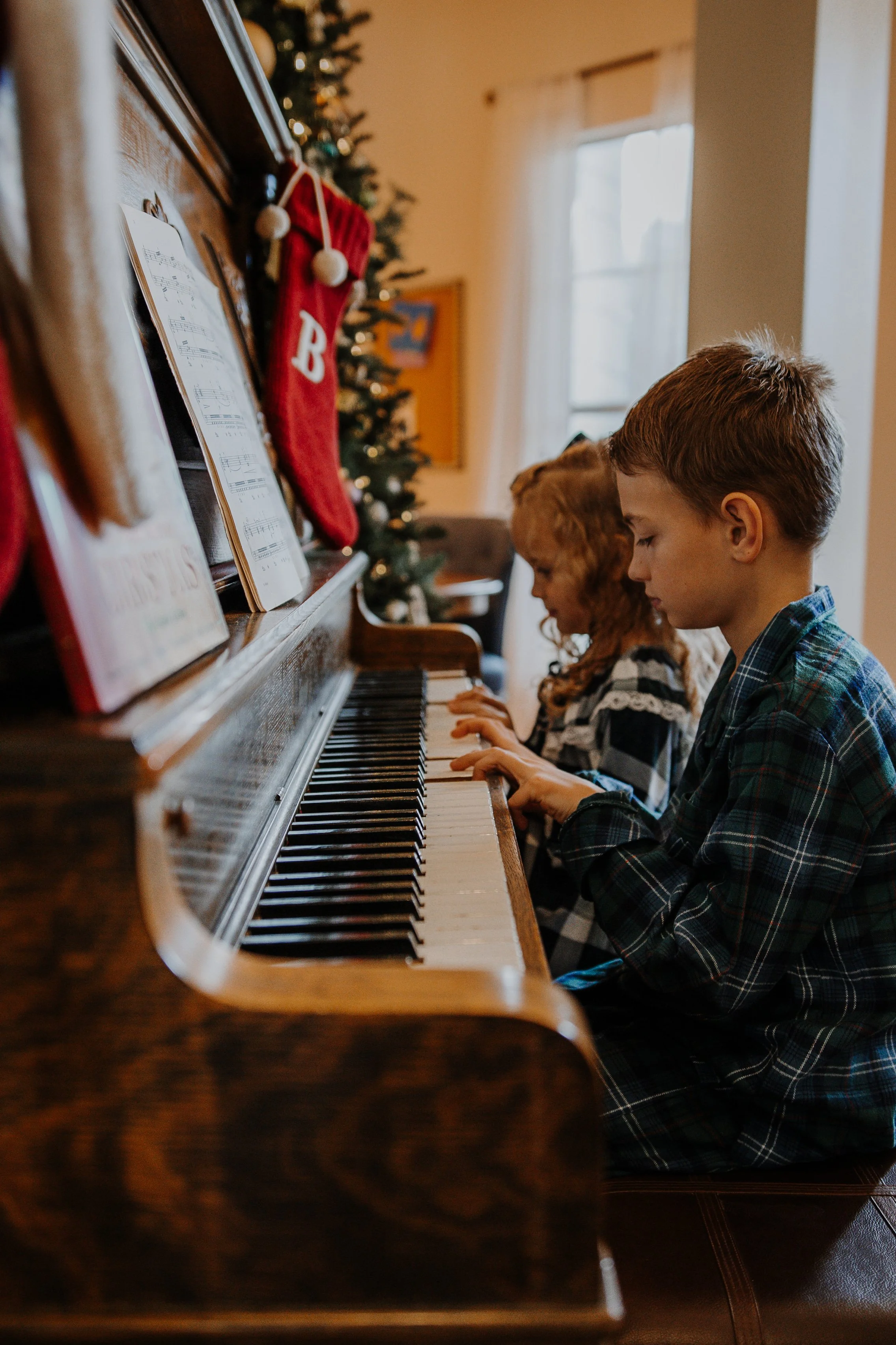 Two children, a boy and a girl, playing piano in a room decorated for Christmas with a Christmas tree, stockings, and music sheets.