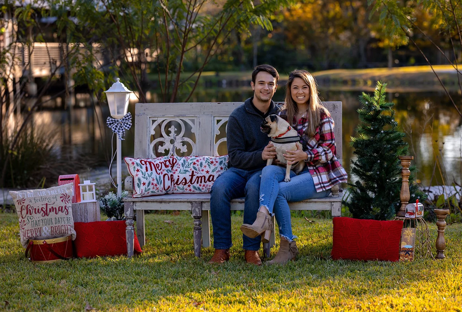 A young couple sitting on a decorative white bench by a lake, holding a dog, surrounded by Christmas decorations including a small Christmas tree, red and white pillows, and holiday-themed items. The scene is set outdoors during daylight.