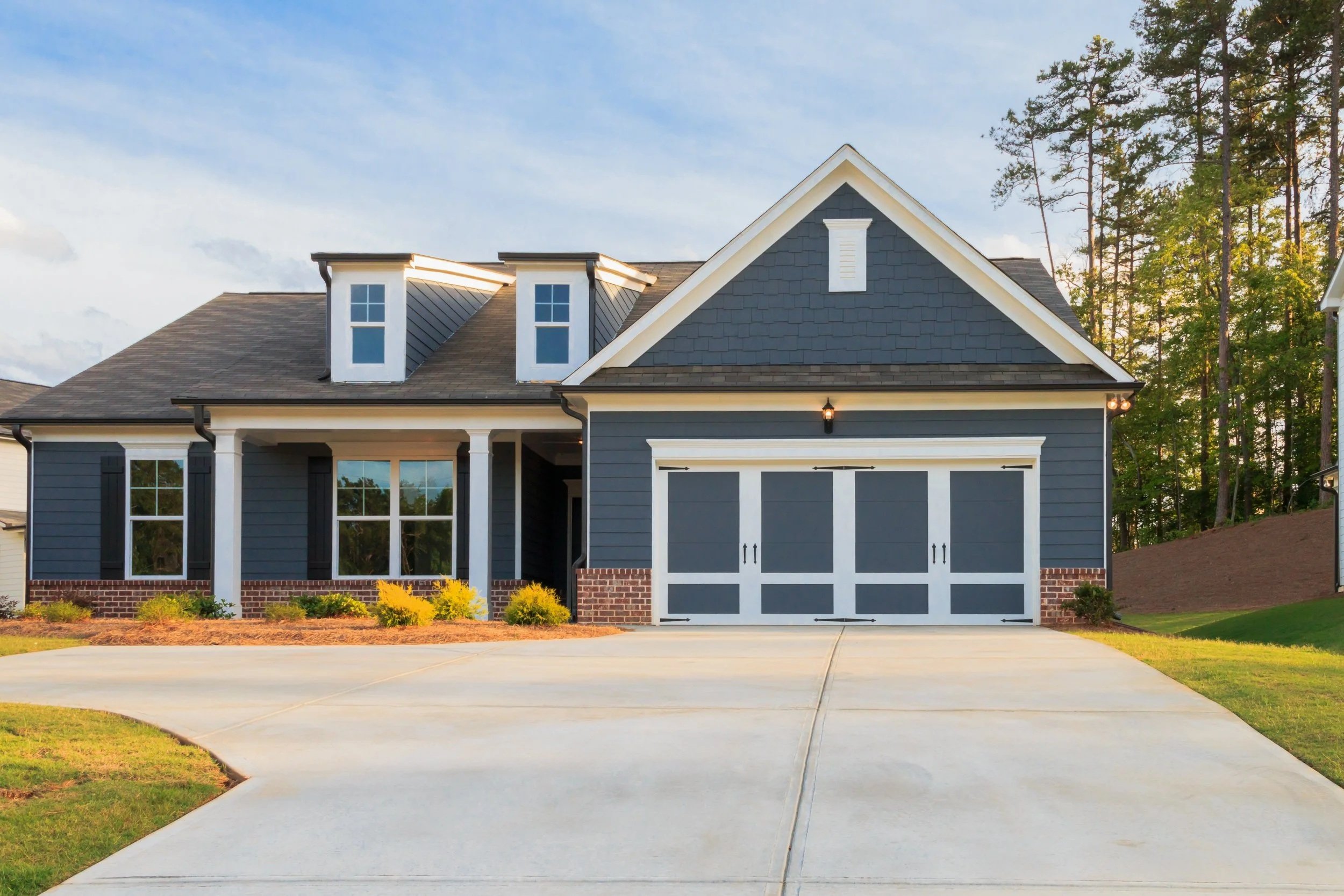 Front view of a modern house with a gable roof, attached garage, and a well-maintained lawn, situated next to a wooded area.
