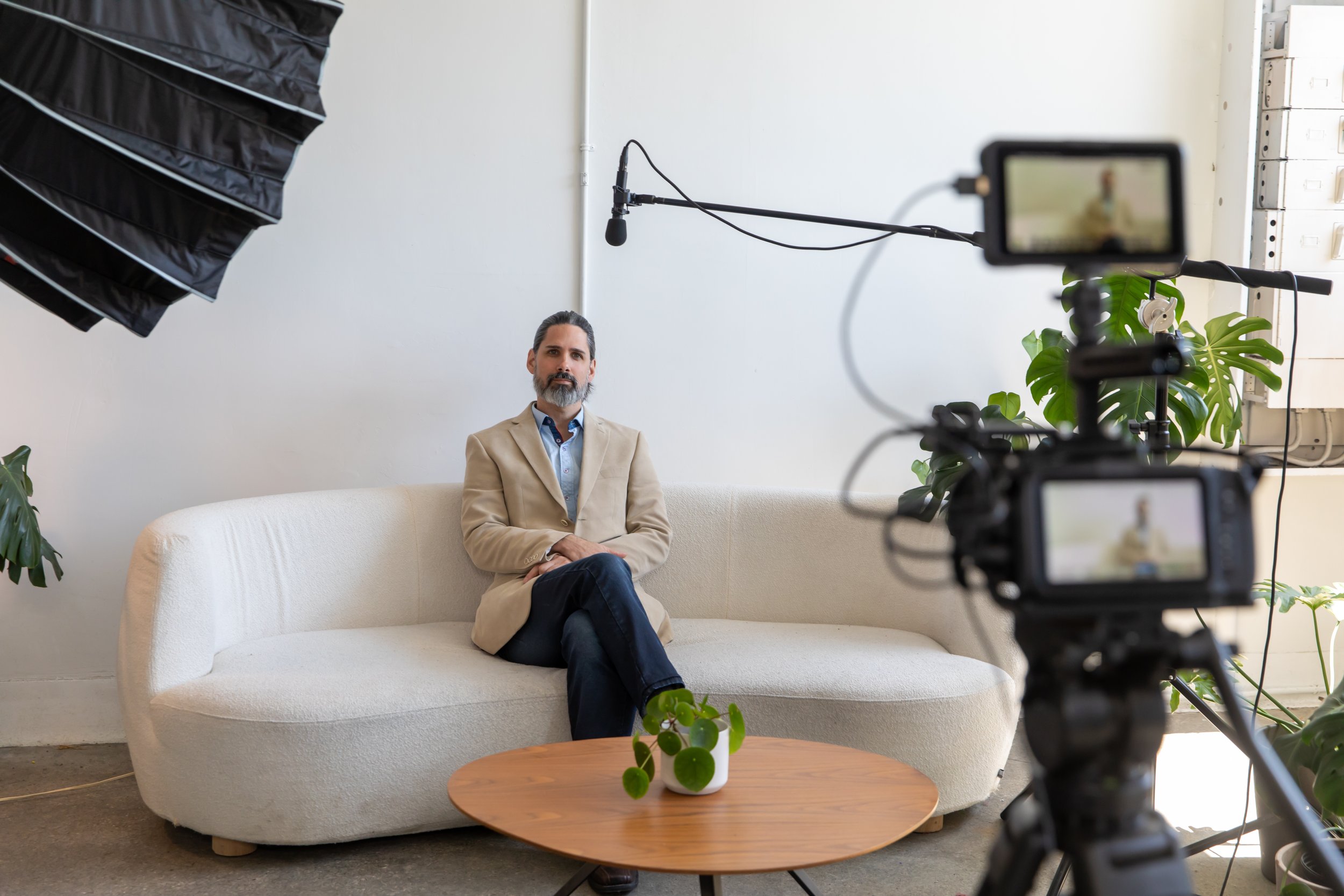 A man with a beard and mustache, wearing a beige blazer and blue shirt, sitting on a white sofa in a well-lit room with white walls, plants, and a small wooden table in front of him. A camera and lighting equipment are set up for filming.