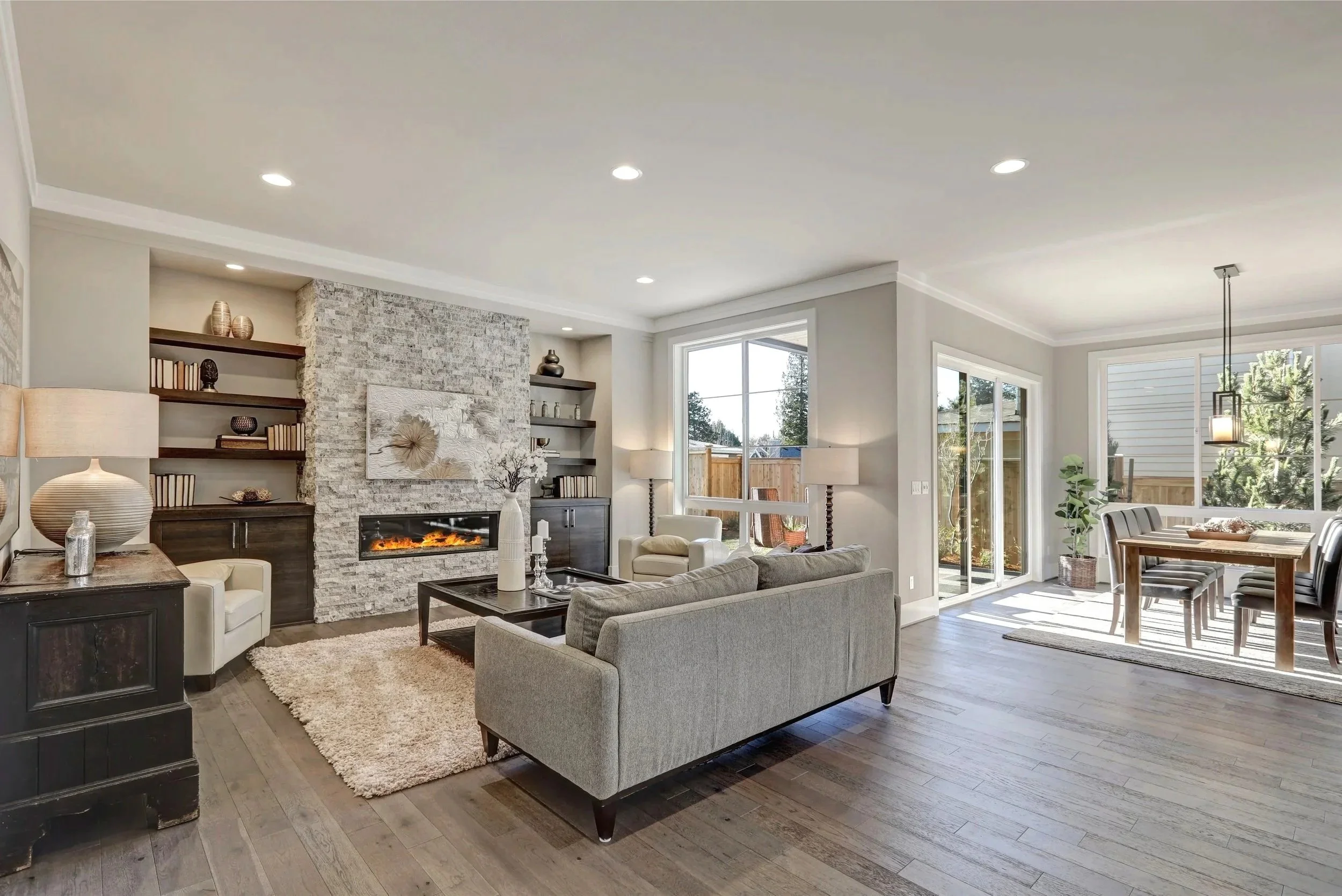 Open-concept living room and dining area with large windows, a stone fireplace, modern furniture, and natural wood flooring.