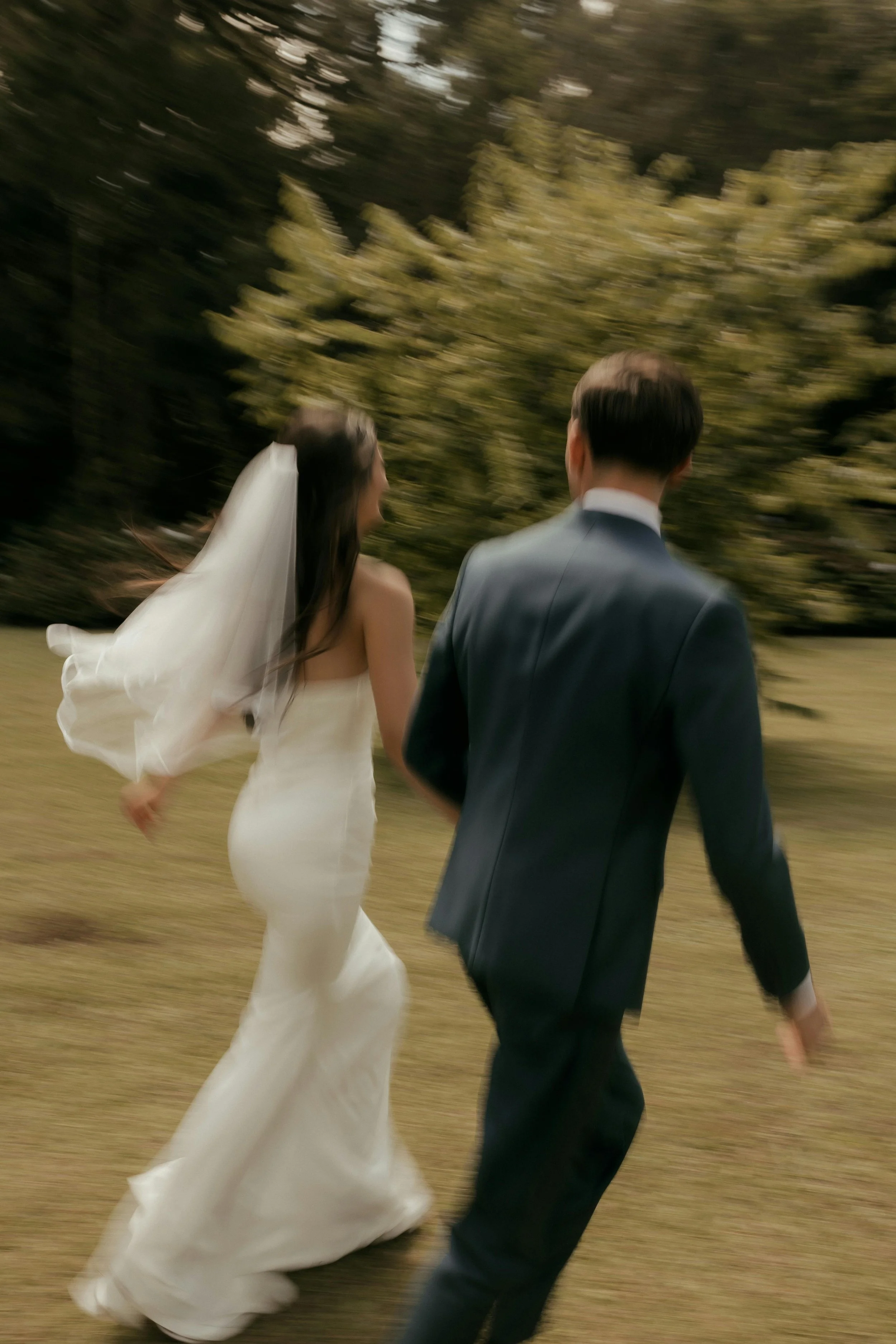 A bride and groom walking outdoors, the bride in a white wedding dress and veil, the groom in a dark suit, with trees and greenery in the background.