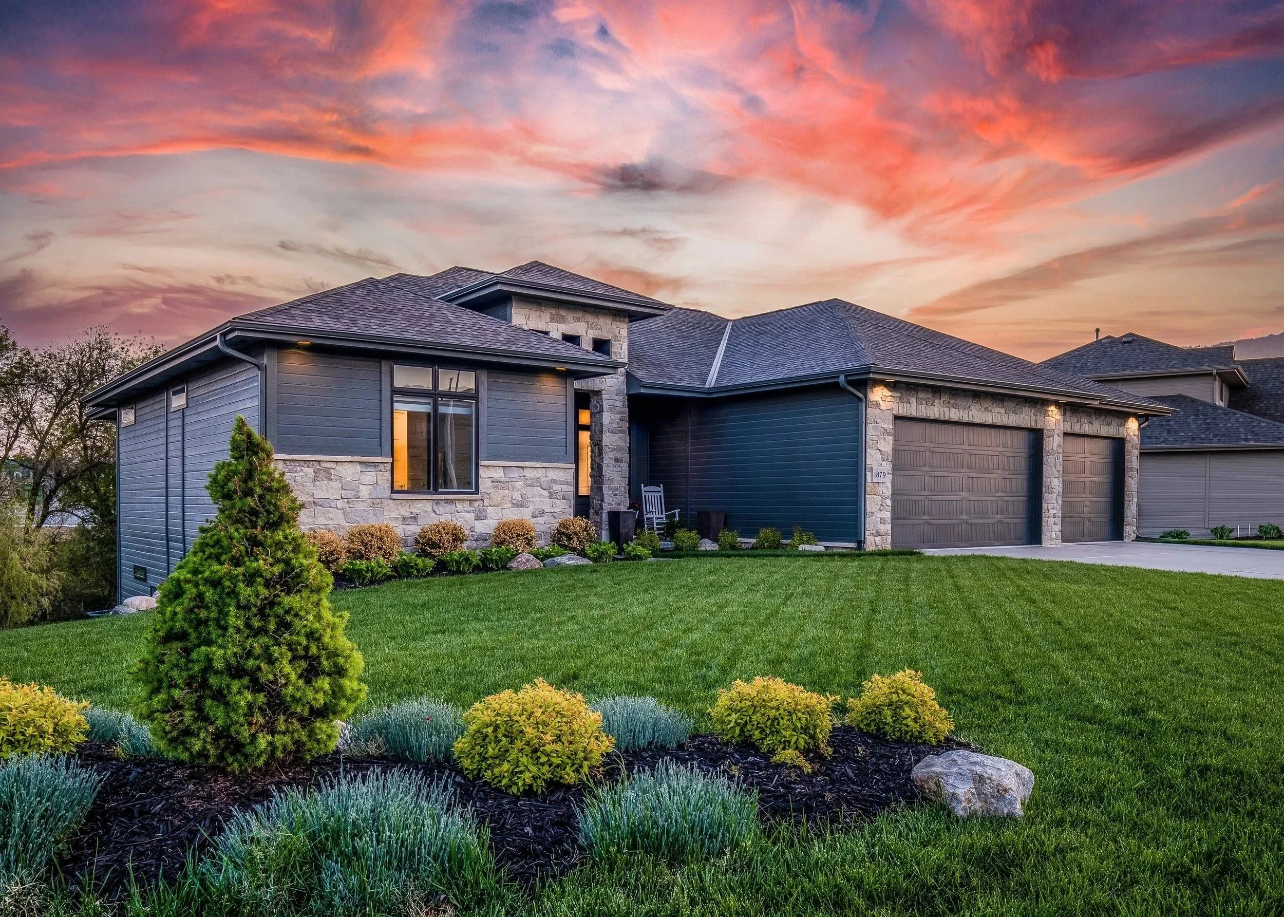 A modern house with a gray and stone facade, surrounded by a well-maintained green lawn and landscaped garden with shrubs and small plants, under a colorful sunset sky with pink, orange, and purple clouds.