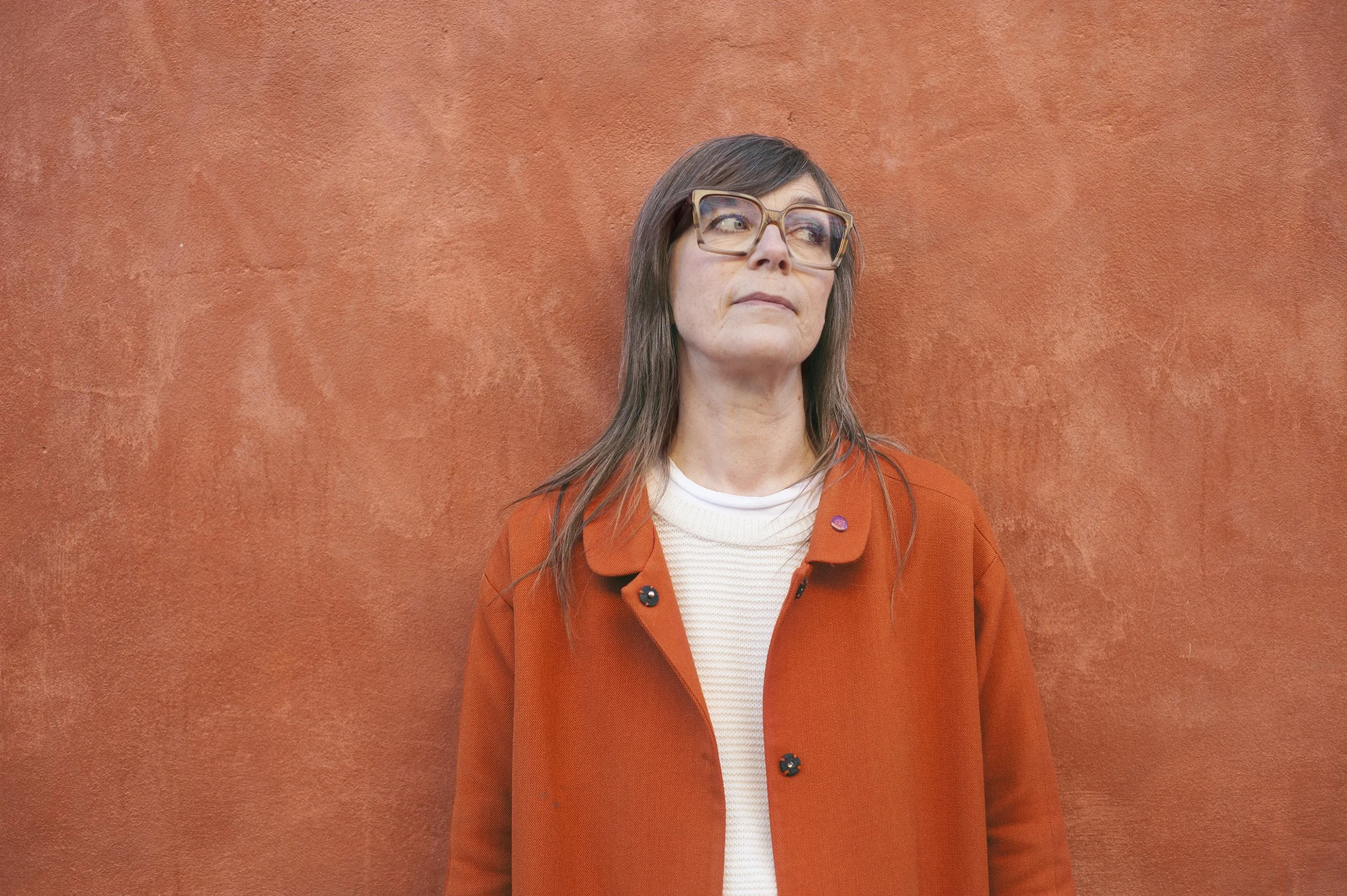 Halvbild på Karin Lönnå i vit tröja och orange kappa. Hon står lutad mot en rödbrun putsad vägg. Foto: Kerstin Skjaervold