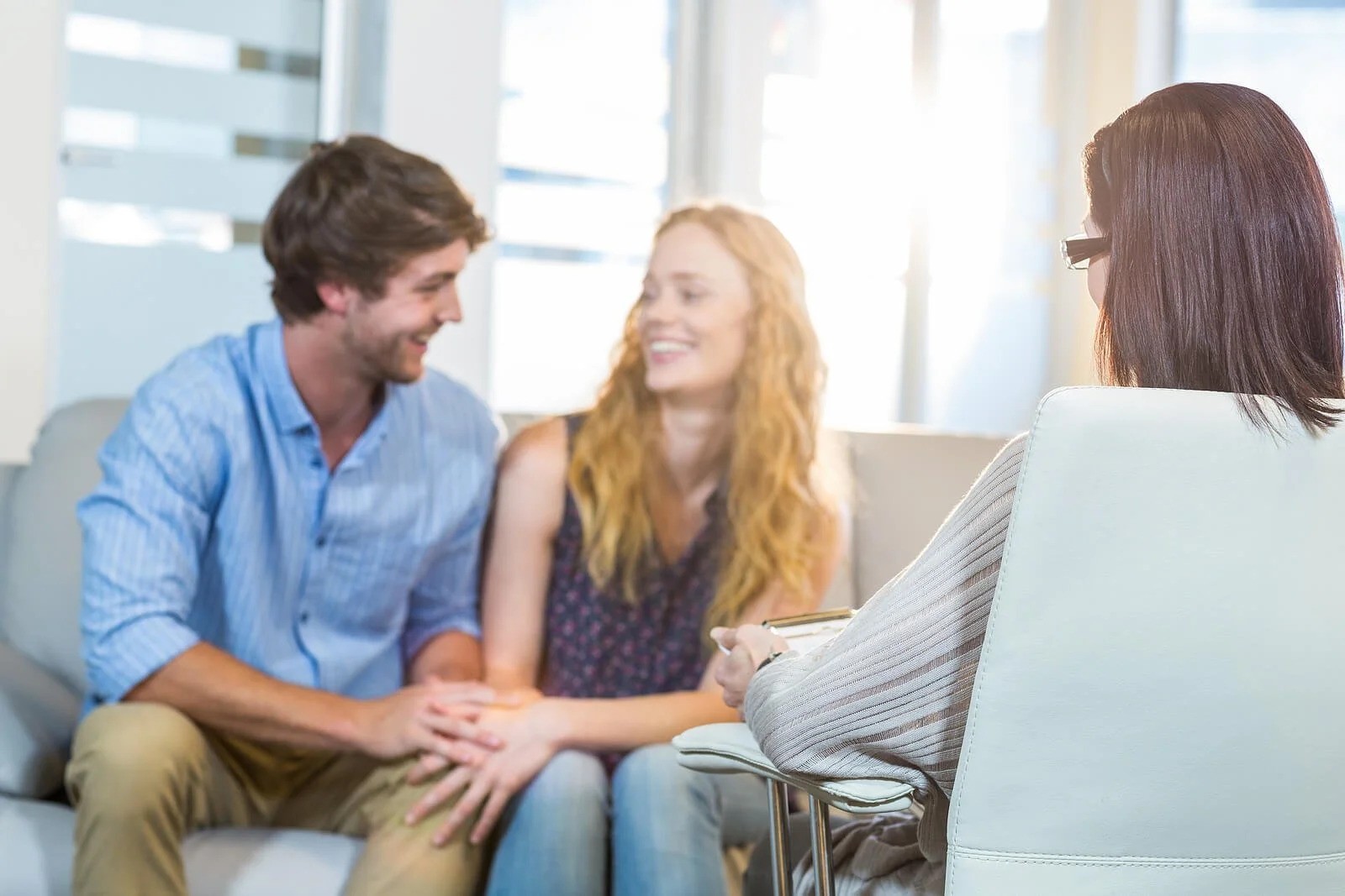 Smiling engaged couple sitting on couch talking with female counselor holding clipboard. Address finances, conflict, and expectations before you say "I do" with premarital counseling in Tampa, FL.