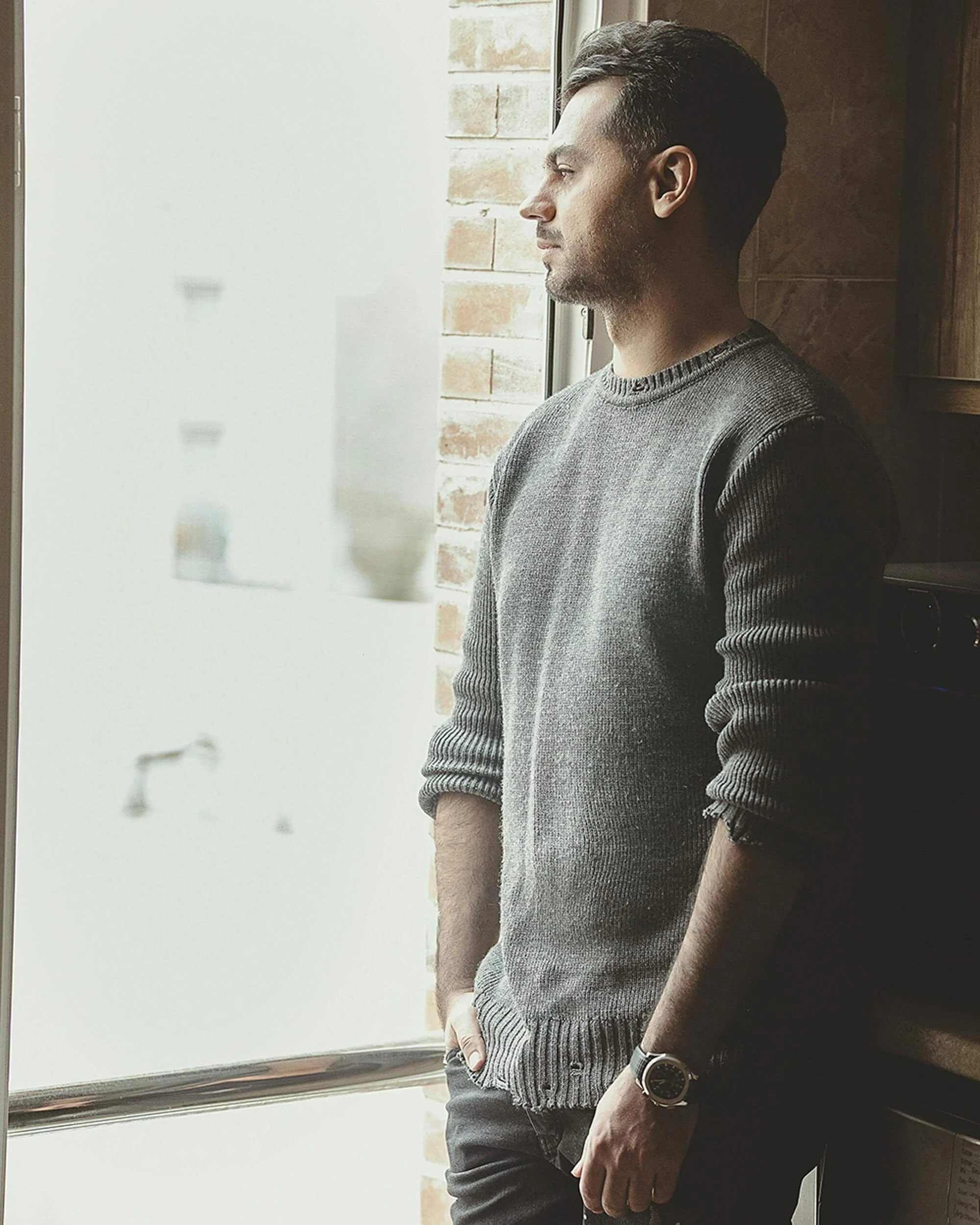 Man in gray sweater standing by window looking contemplative in natural light. Learn how Christians are finding relief from stuck trauma through EMDR therapy in Tampa, FL.