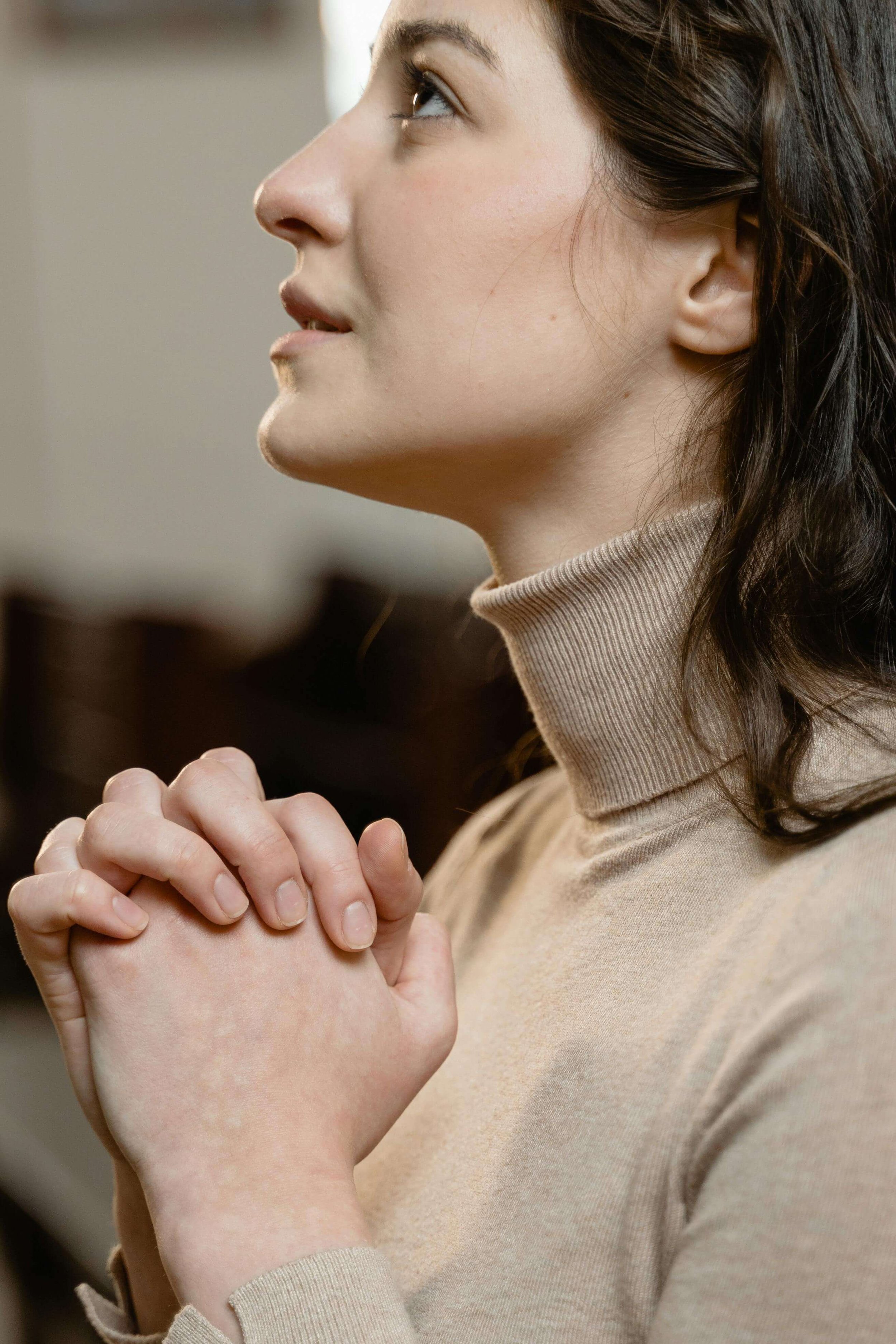 Woman in beige turtleneck with hands clasped in prayer looking upward. When faith and grief coexist, find compassionate support through Christian counseling for grief in Tampa, FL.