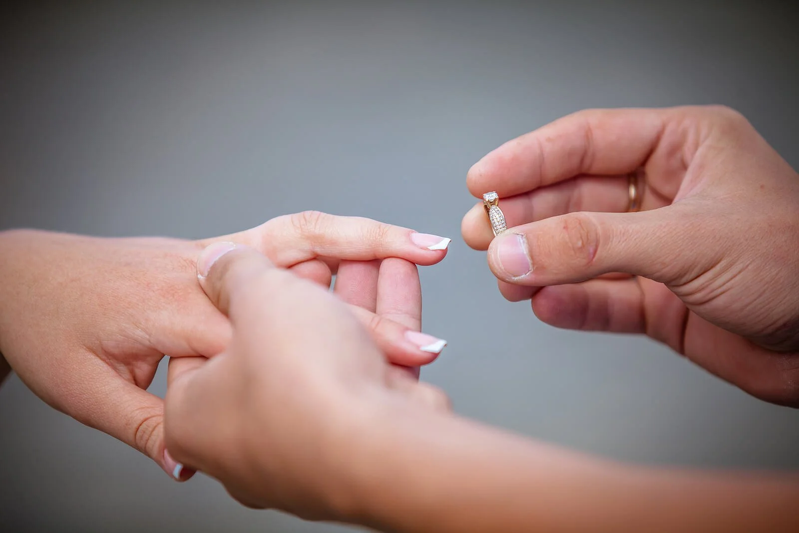 Hands of engaged couple with engagement ring being placed on finger against gray background. Prepare for marriage, not just the wedding, with SYMBIS-based premarital counseling in Tampa, FL.