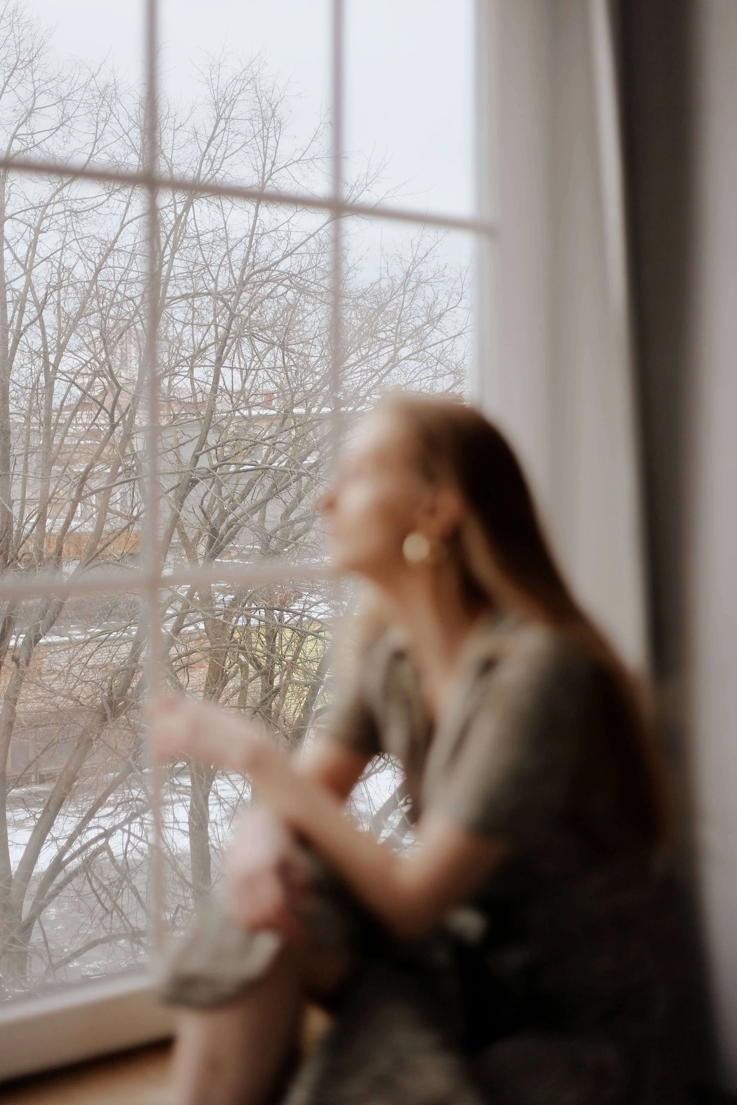 A blurred woman sitting by a large window looking outside at bare winter trees. Healing from the past is possible and EMDR therapy in Tampa, FL gives you a way forward without having to relive every detail.
