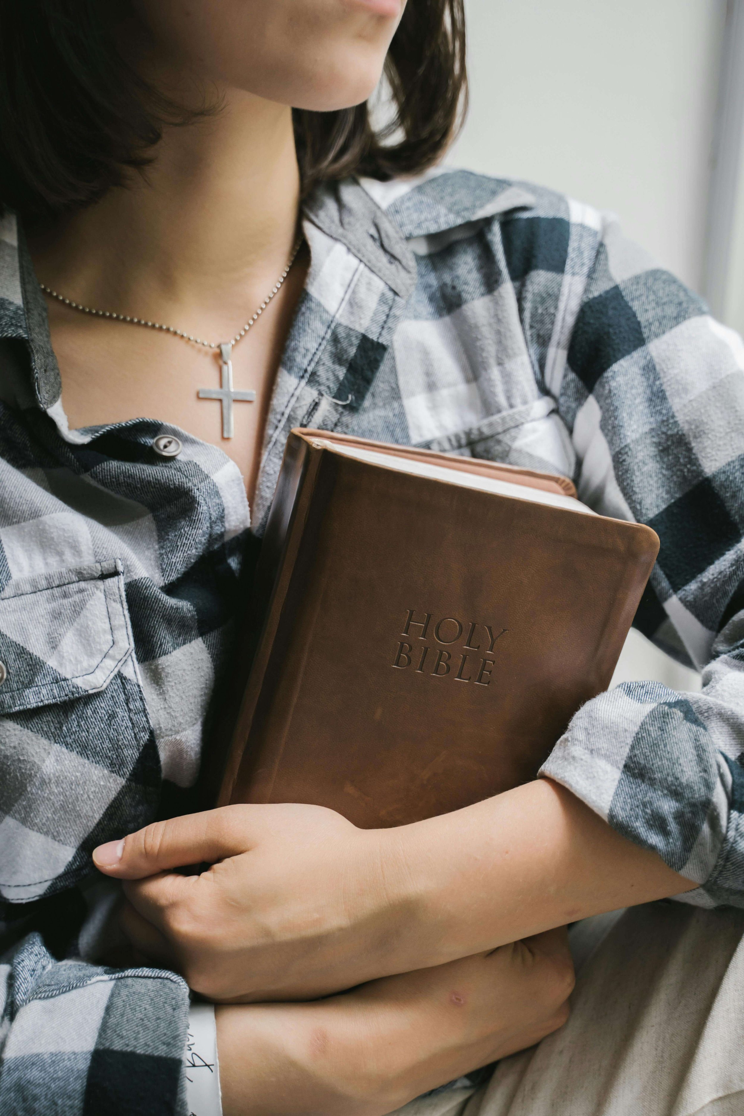 A woman wearing a cross necklace and plaid shirt holds a Holy Bible close to her chest. Explore how Scripture deepens your healing journey through Christian Counseling in Tampa, FL.
