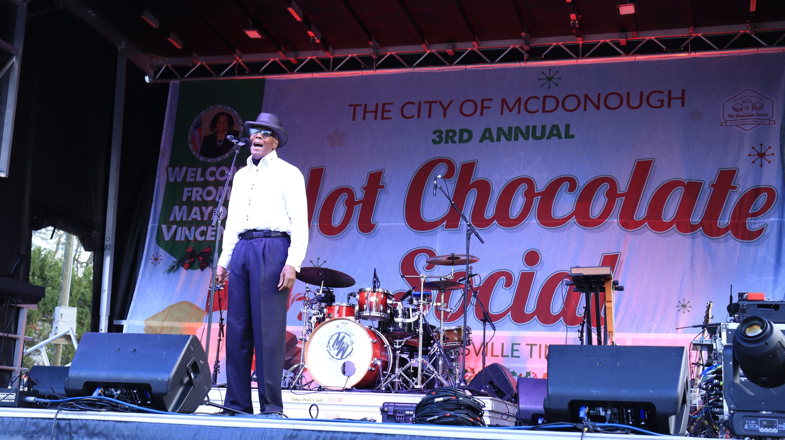 A man wearing a black hat, sunglasses, a white shirt, and dark pants standing on a stage with a microphone. Behind him, there is a backdrop that reads 'The City of McDonough 3rd Annual Hot Chocolate Festival.' On the stage, there is a drum set and so