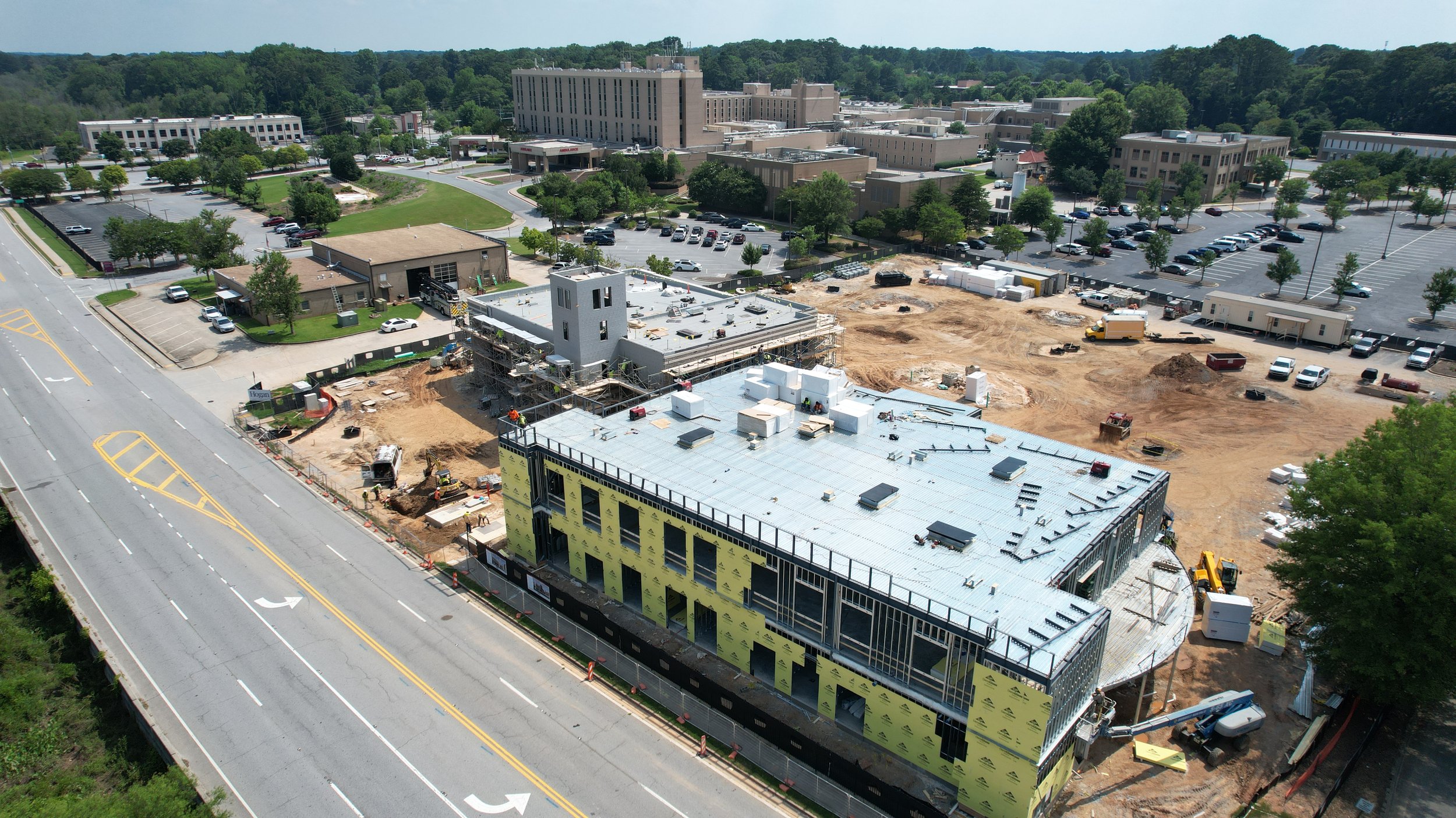 An aerial view of a construction site with a building in progress, surrounded by parking lots and greenery in the background.