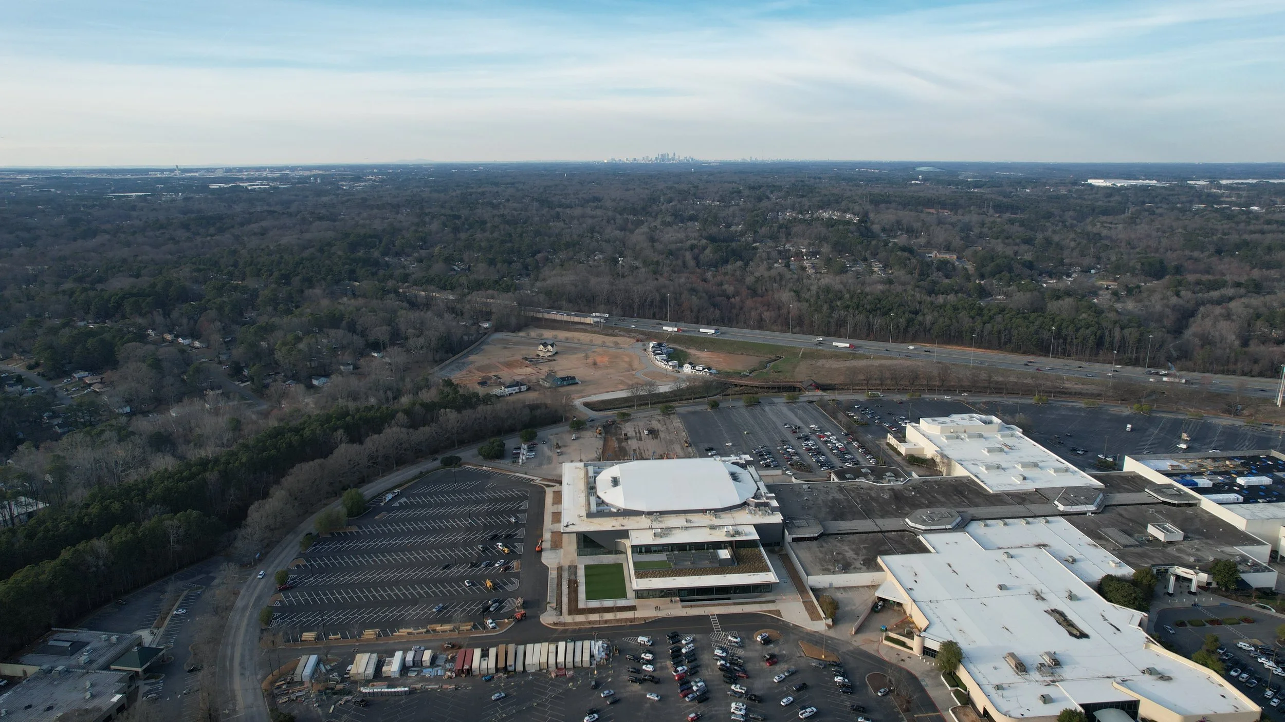 An aerial view of a commercial area with a large parking lot, a shopping mall or office building with a white roof, and nearby highways and a wooded area in the background.