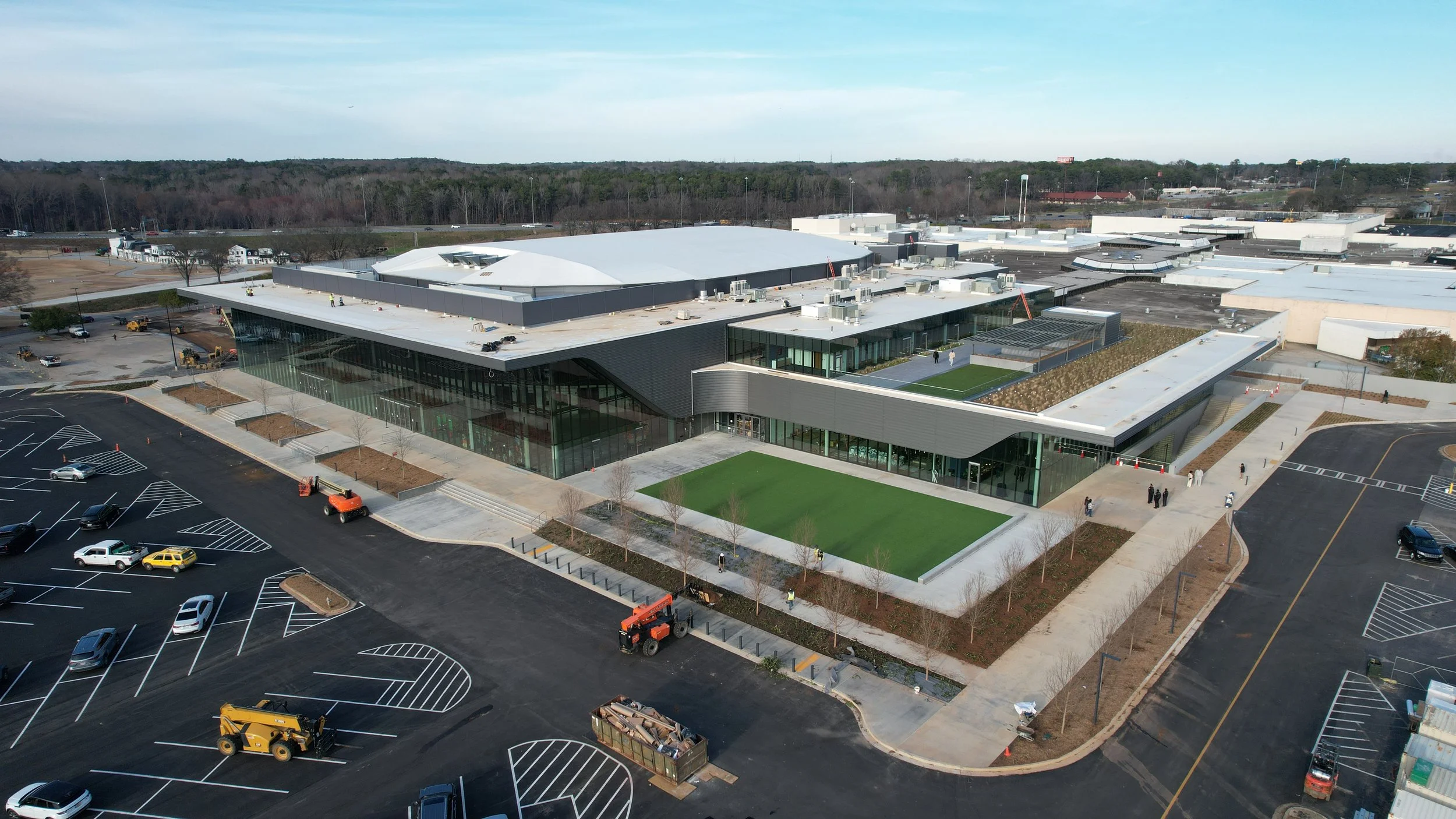An aerial view of a large modern building with a parking lot in the foreground and a wooded area in the background.