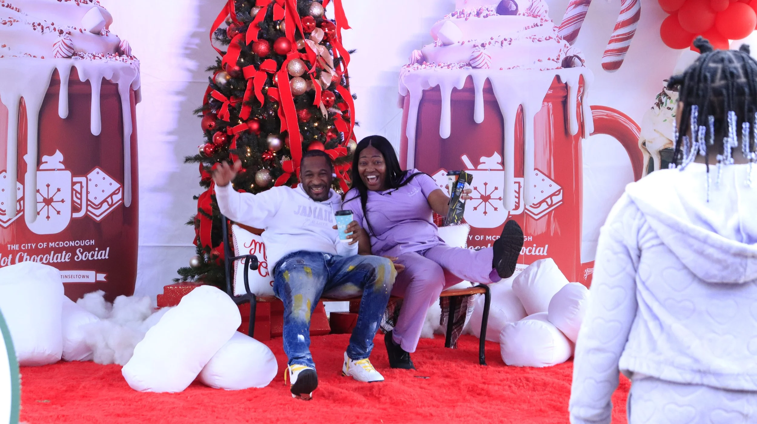 Two people sitting on a bench, smiling and having fun, in front of a Christmas themed backdrop featuring a decorated Christmas tree and large holiday-themed images of hot chocolate mugs and candy Canes.