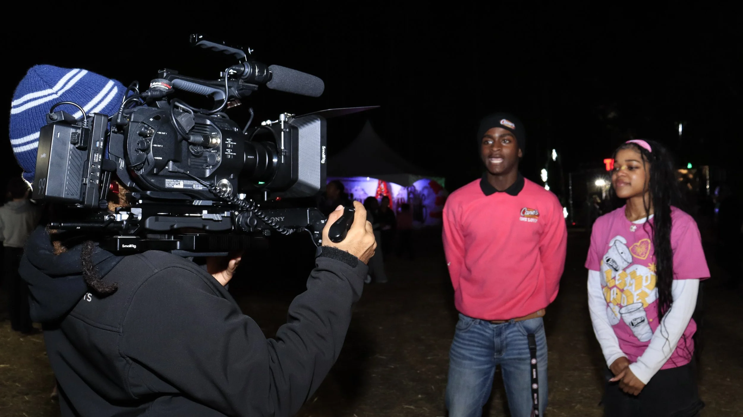 A person operating a professional video camera at night, filming two individuals who are standing and talking outdoors, with lights and tents in the background.