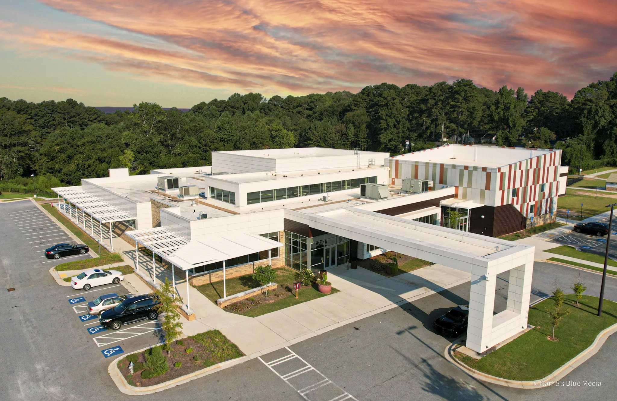 Aerial view of a modern school building with multiple sections, parking lot with some cars, landscaped areas, and a treeline in the background during sunset.