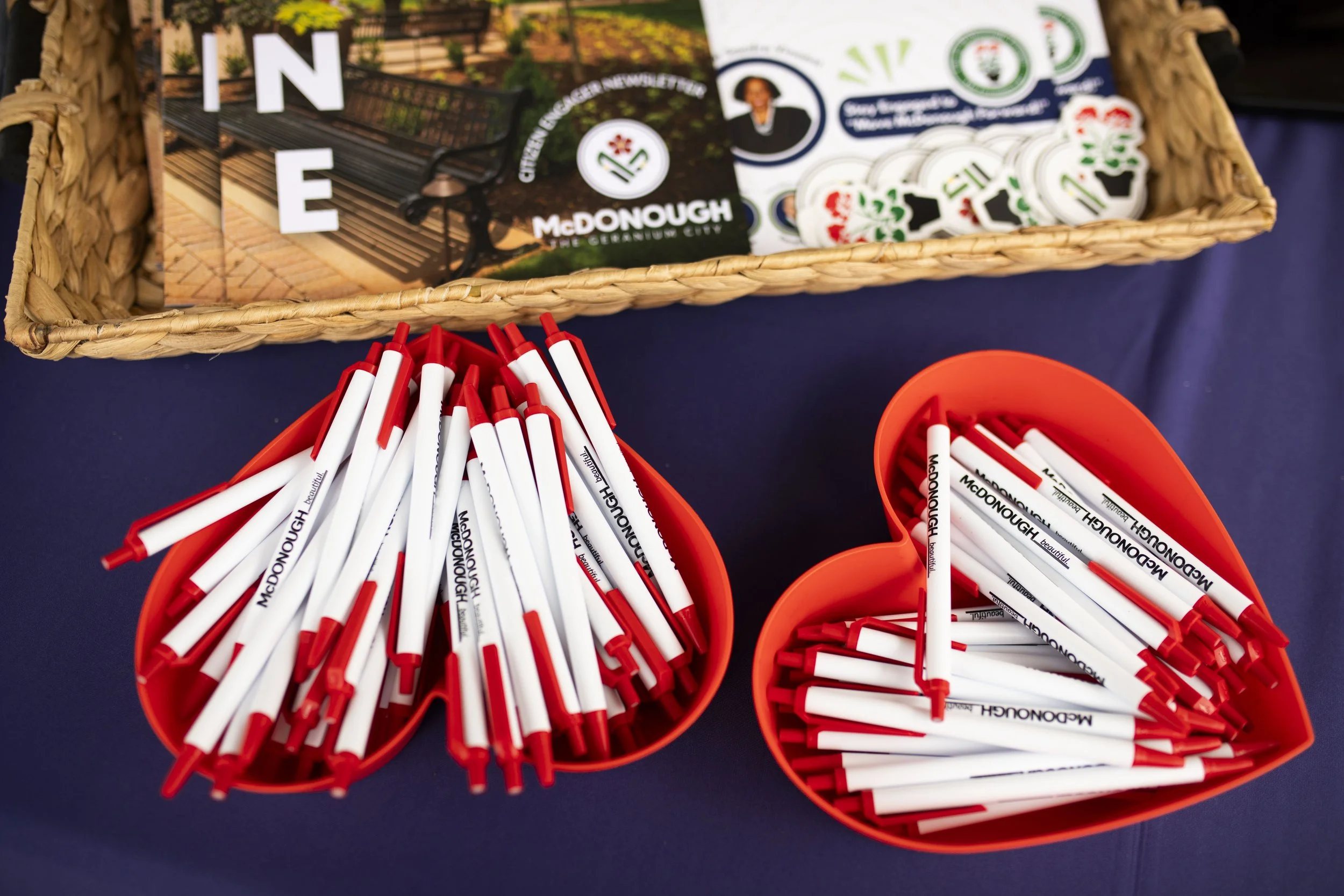 Red and white pens with 'McDonough' printed on them placed in orange heart-shaped containers on a table, with a basket of flyers and stickers nearby promoting McDonough, Georgia.