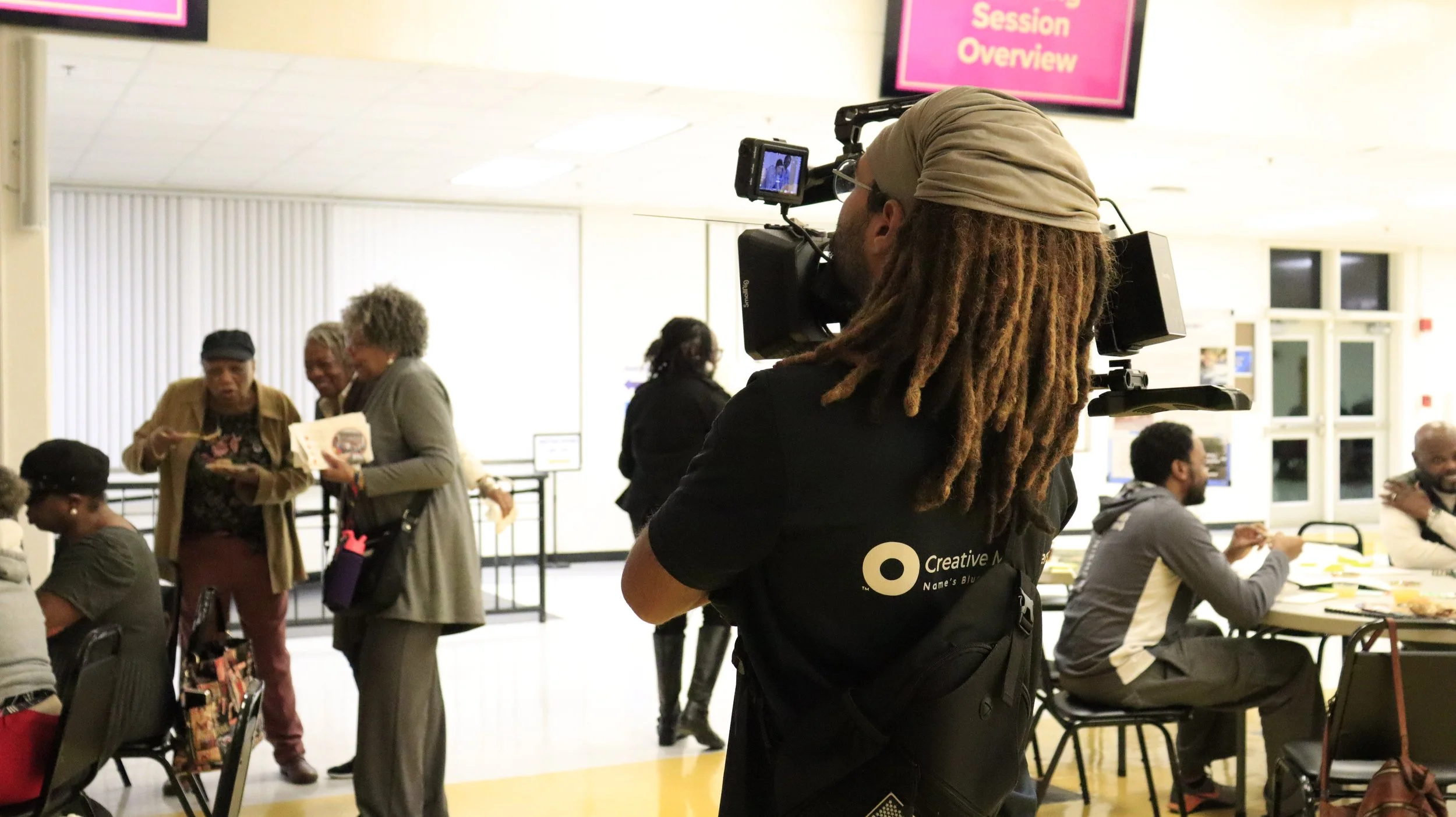 A person with dreadlocks and a beige head wrap using a professional video camera to record a social gathering in a well-lit indoor space. People are standing and sitting around tables, engaging in conversations. A pink sign that reads 'Session Overview' is visible in the background.