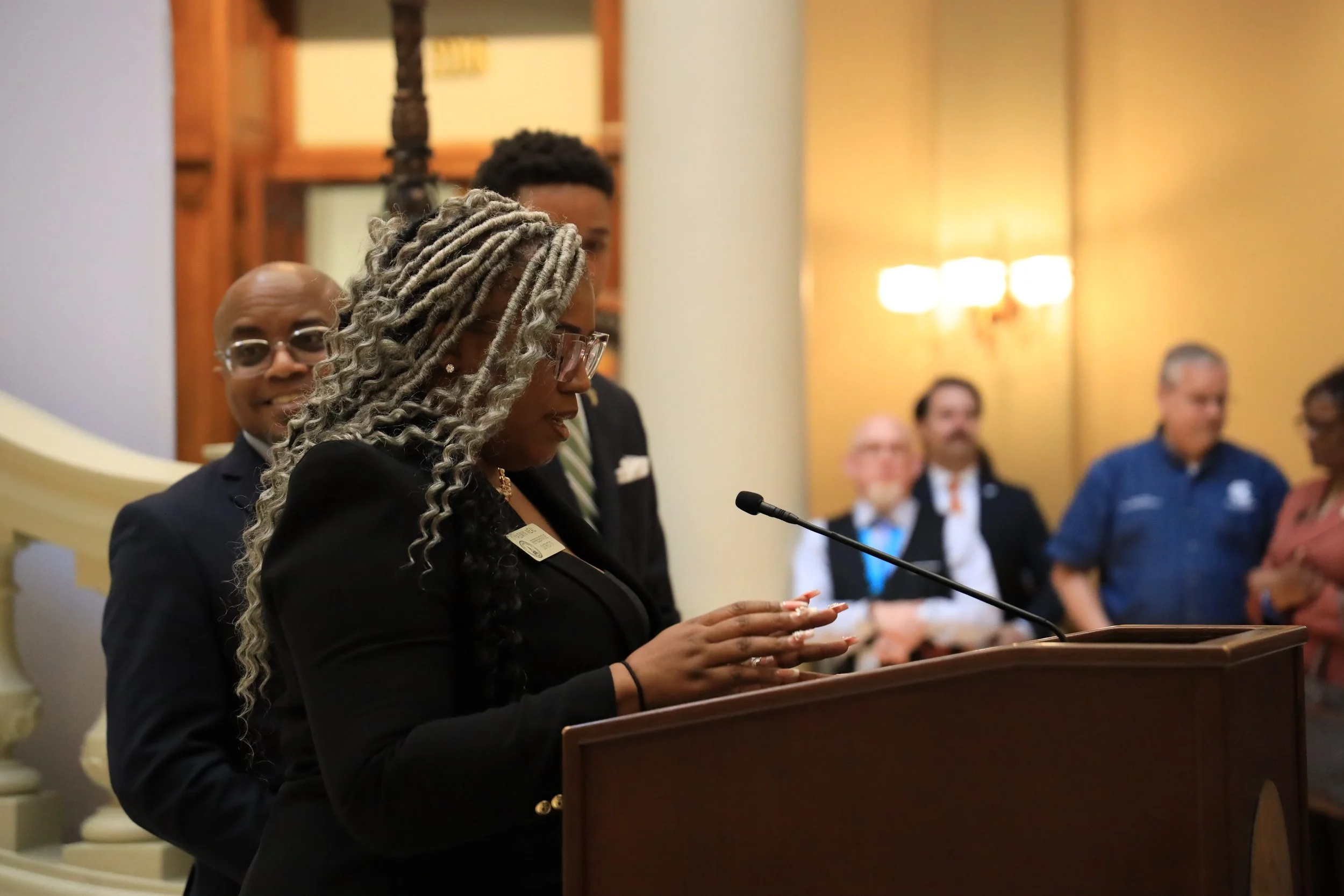 A woman with long, curly gray hair and glasses standing at a podium, speaking into a microphone during a formal event, with several people in business attire standing behind her and a warmly lit room.