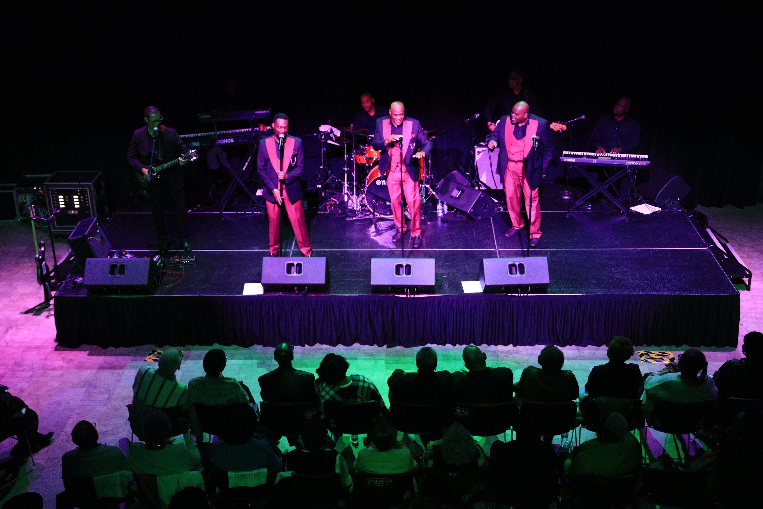 A live music performance on stage with six musicians, including singers and instrumentalists, under colorful stage lighting, in front of an audience seated in a theater or concert hall.