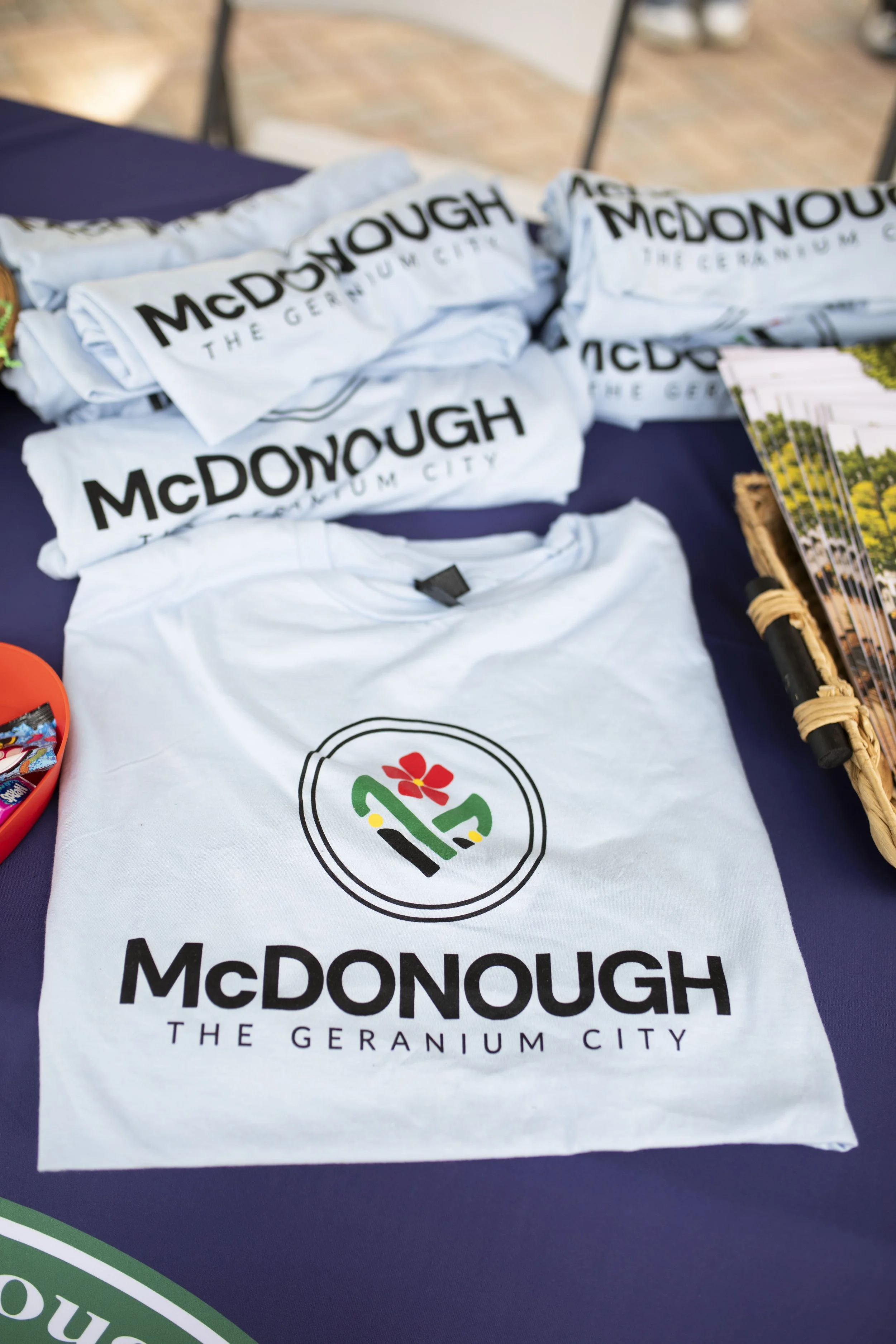 White T-shirt and white cloth bags with 'McDONOUGH THE GERANIUM CITY' logo on a table at an outdoor event, with flyers or brochures and a basket visible on the side.